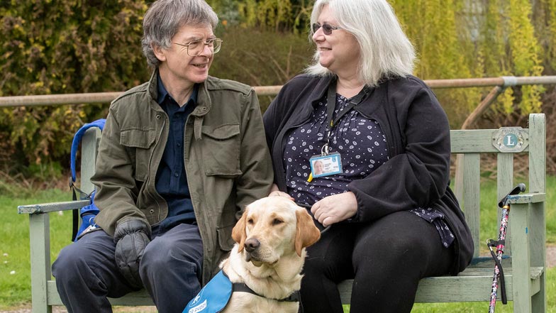 Daisy sitting next to a bench with her puppy walker and Guide Dogs member of staff Daisy sitting next to a bench with her puppy walker and Guide Dogs member of staff
