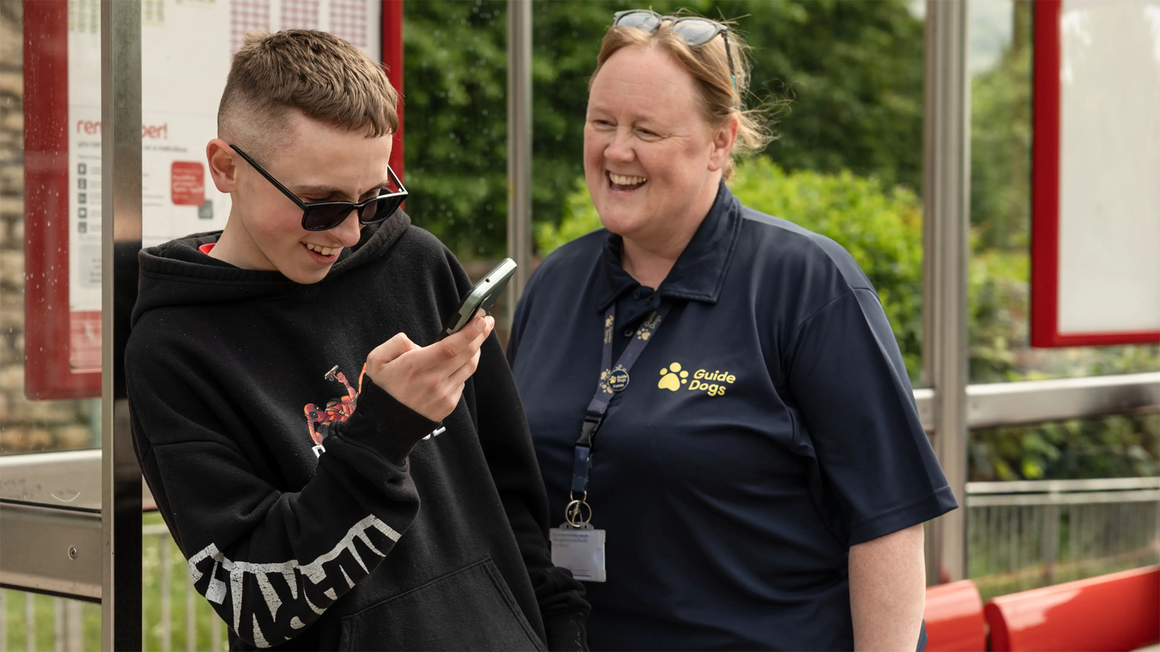 Rhys, a young person with vision impairment, uses his phone at a bus stop. His Habilitation Specialist Kim stands next to him.
