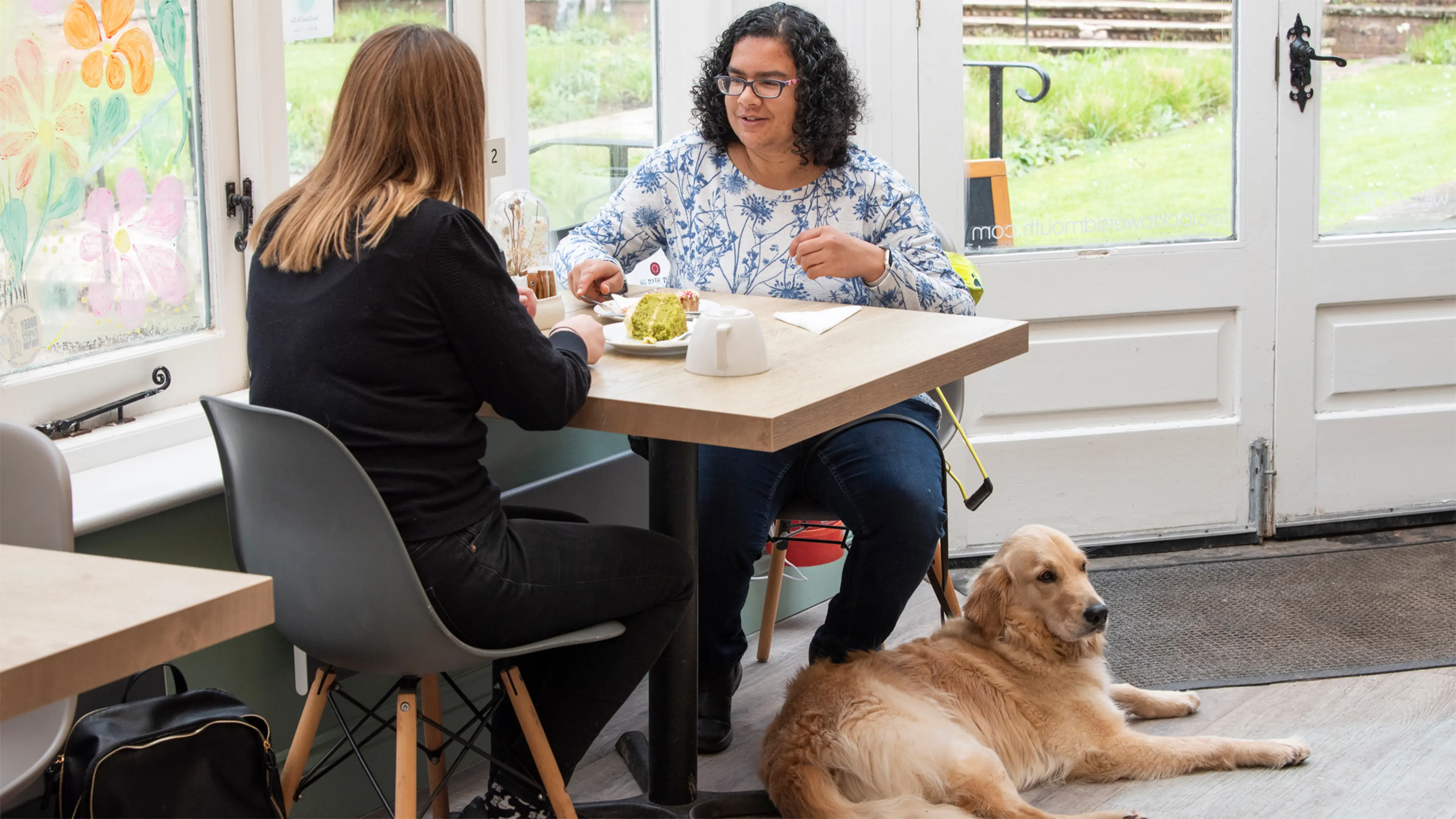 Guide dog owner Lena sits at a cafe table with a friend to have tea and cake. Her guide dog Alex lies at her feet. 