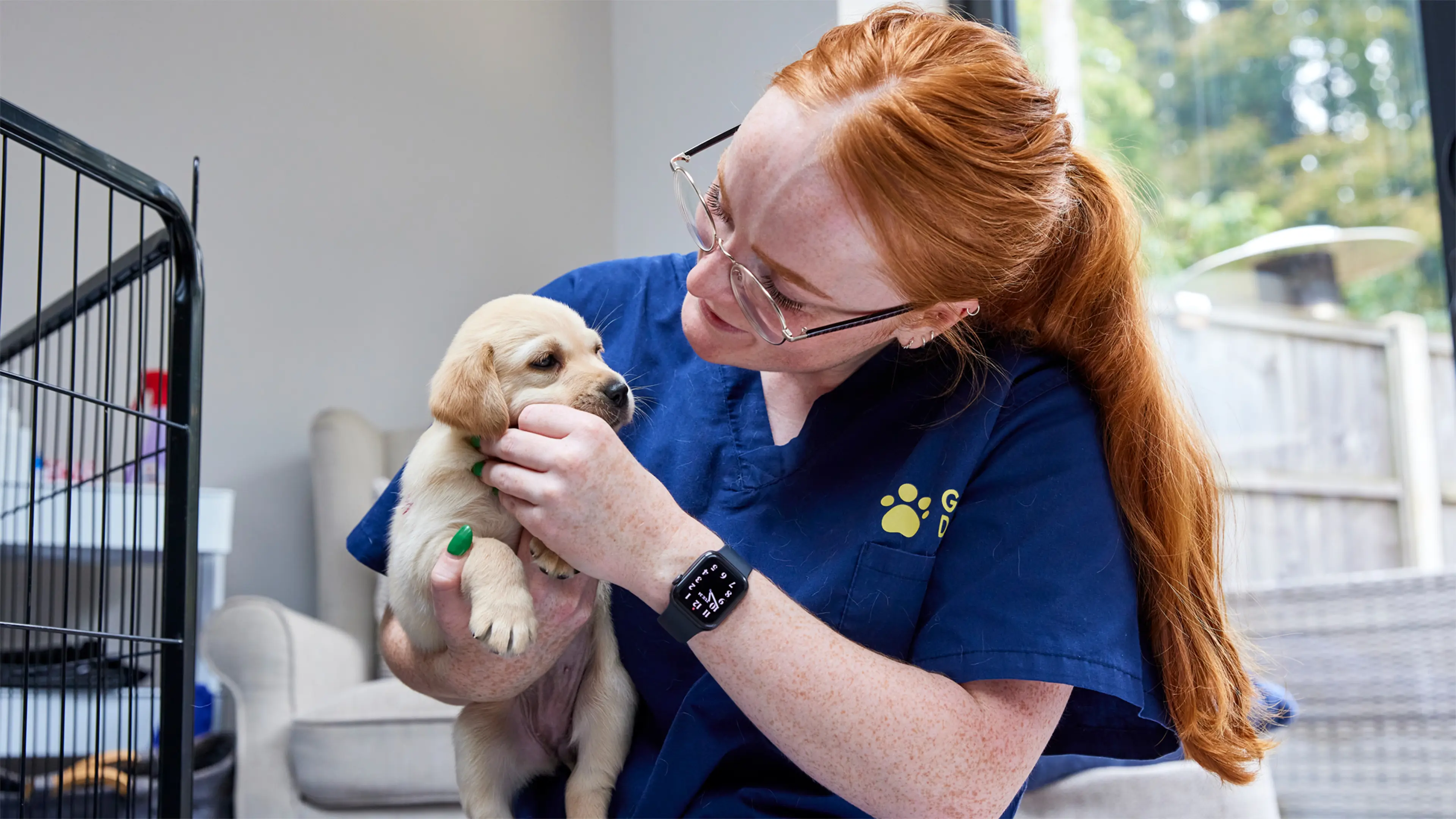 A woman in Guide Dogs scrubs holds a small yellow Labrador puppy as she checks it over at the home of a Breeding Dog Volunteer.