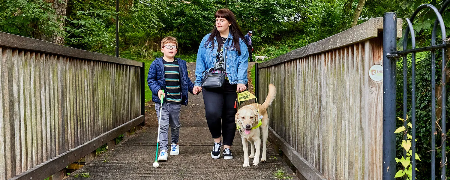 Guide Dog owner Lorna walks across a wooden bridge with her guide dog and son who is using a cane.