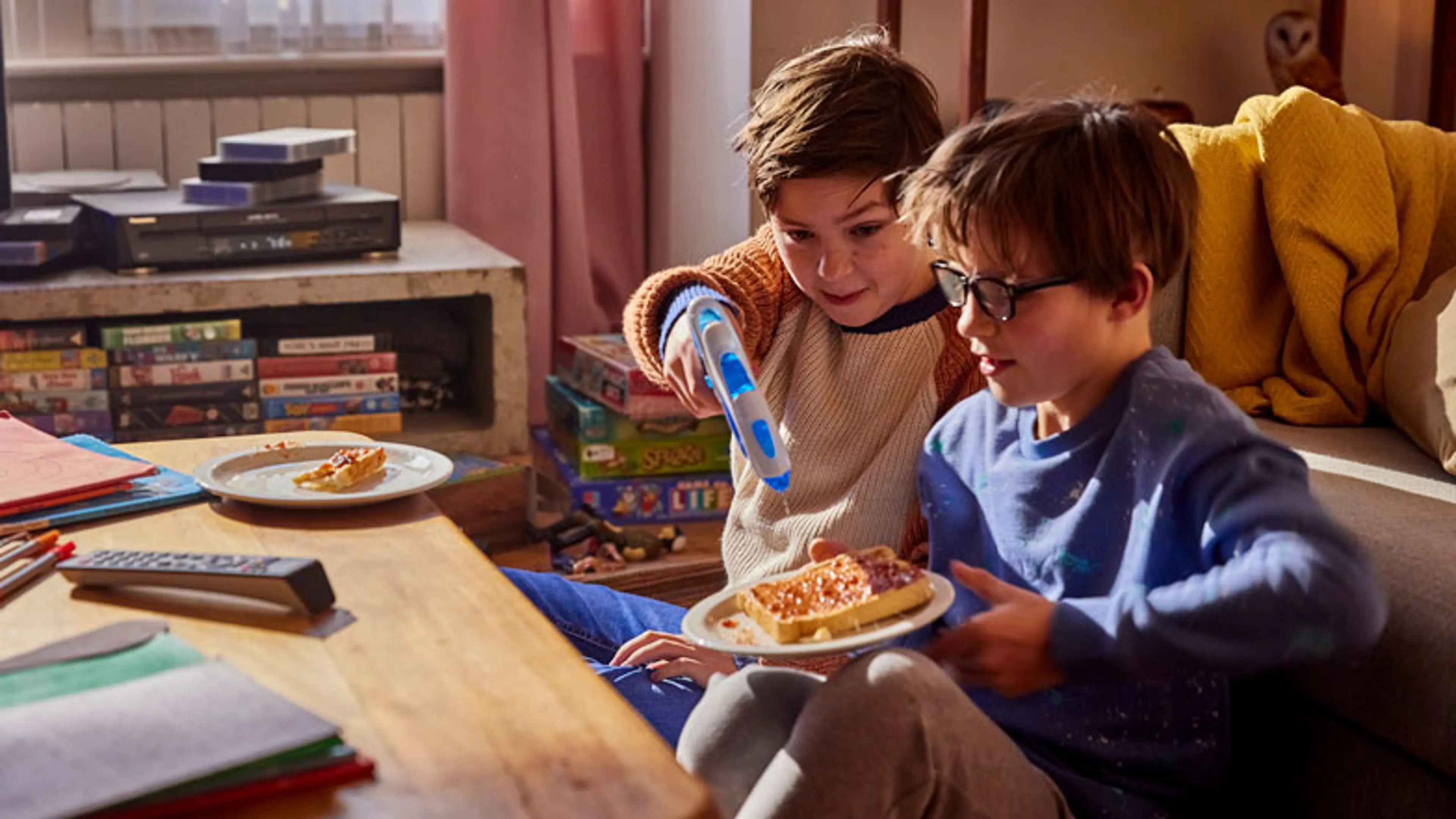 Two young brothers sit on the floor of the living room. One of them holds a piece of toast, and the other a water pistol. They are both laughing.