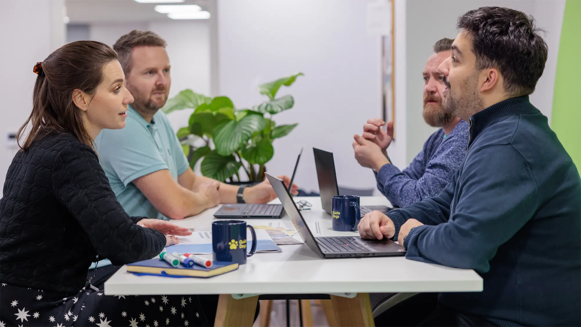 Four members of Guide Dogs staff sit round a table with laptops and notebooks for a meeting. 