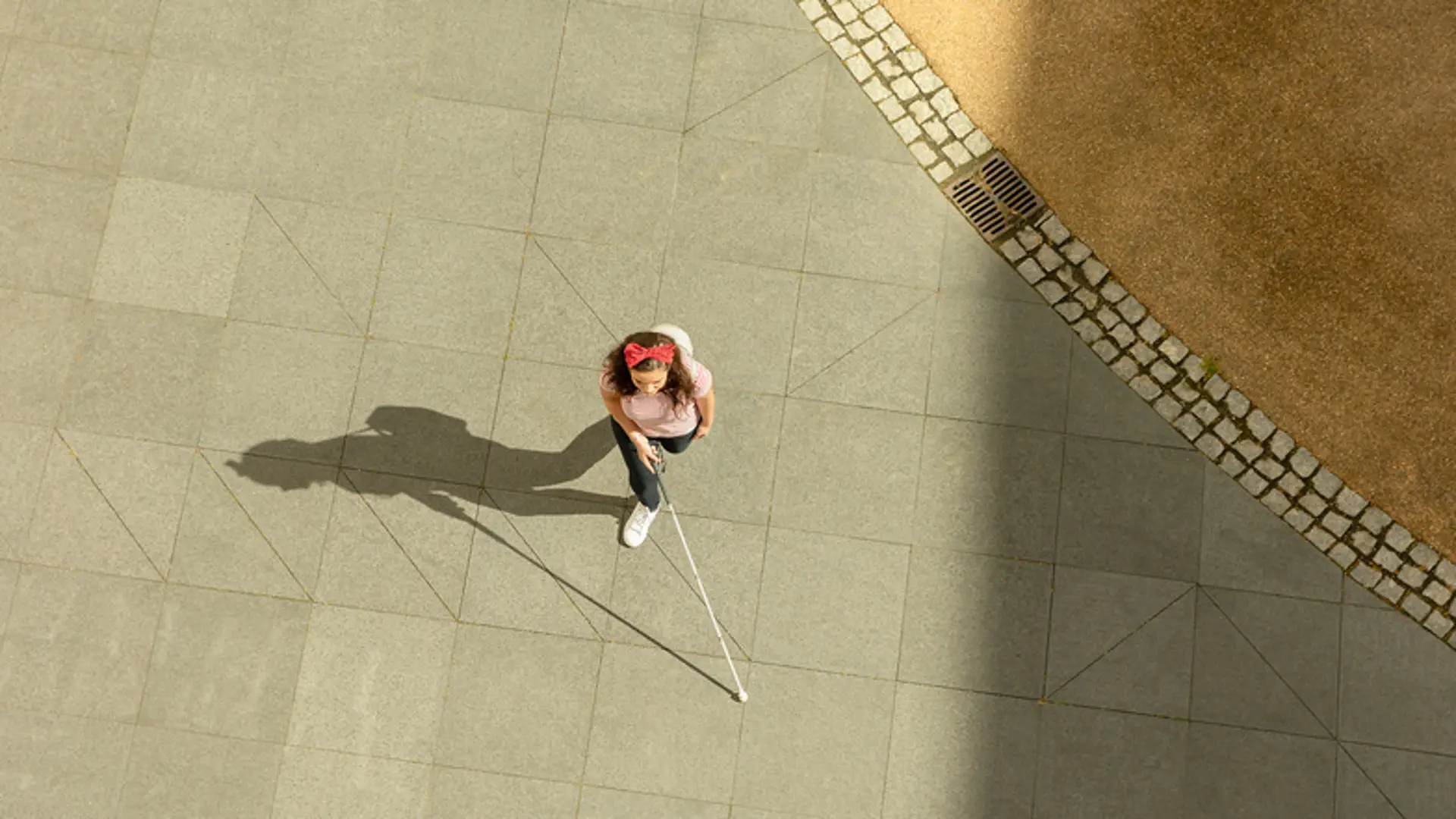 An aerial shot of a woman walking across a courtyard with a long cane. 