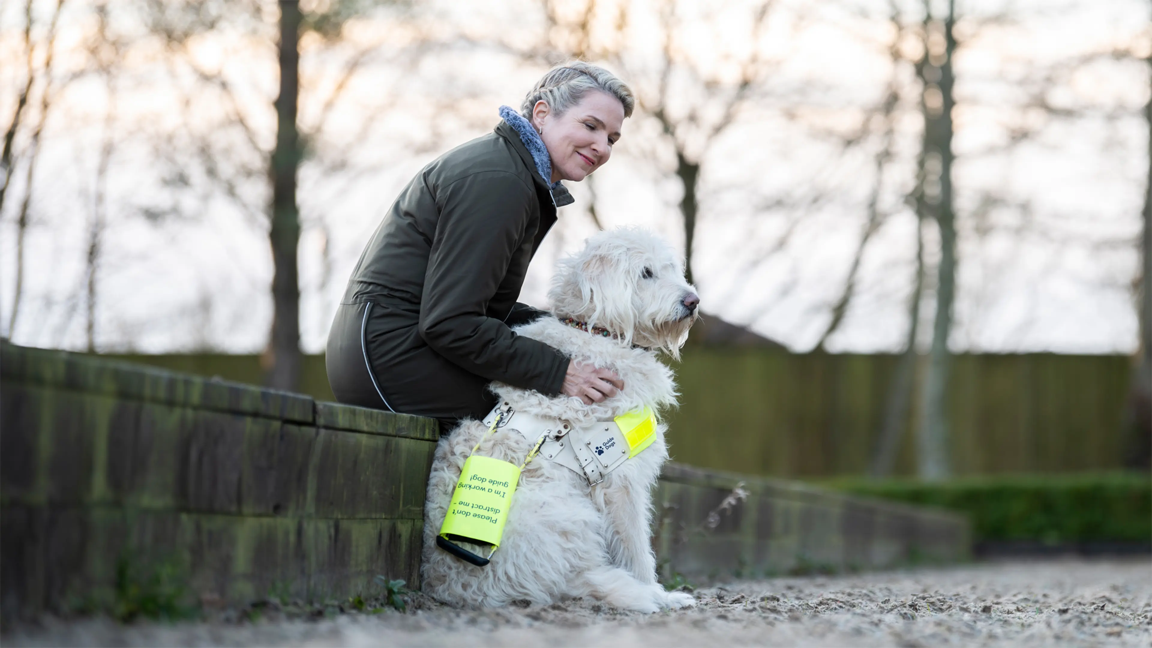 Guide dog owner Verity sits on a low wall next to her guide dog Luna, who wears the Guide Dogs' harness and flash.