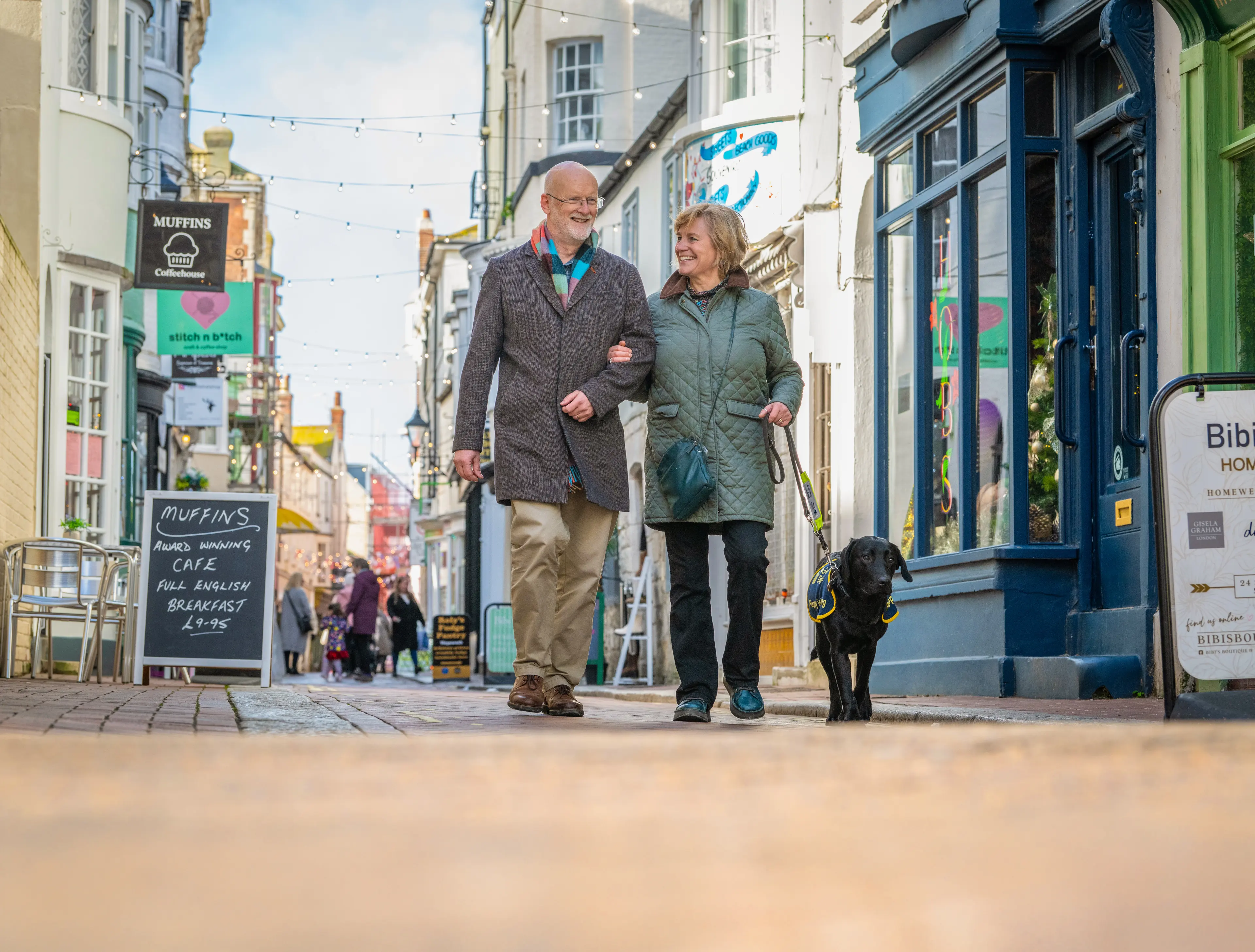 Volunteer puppy raisers walk along a high street smiling with a guide dog puppy