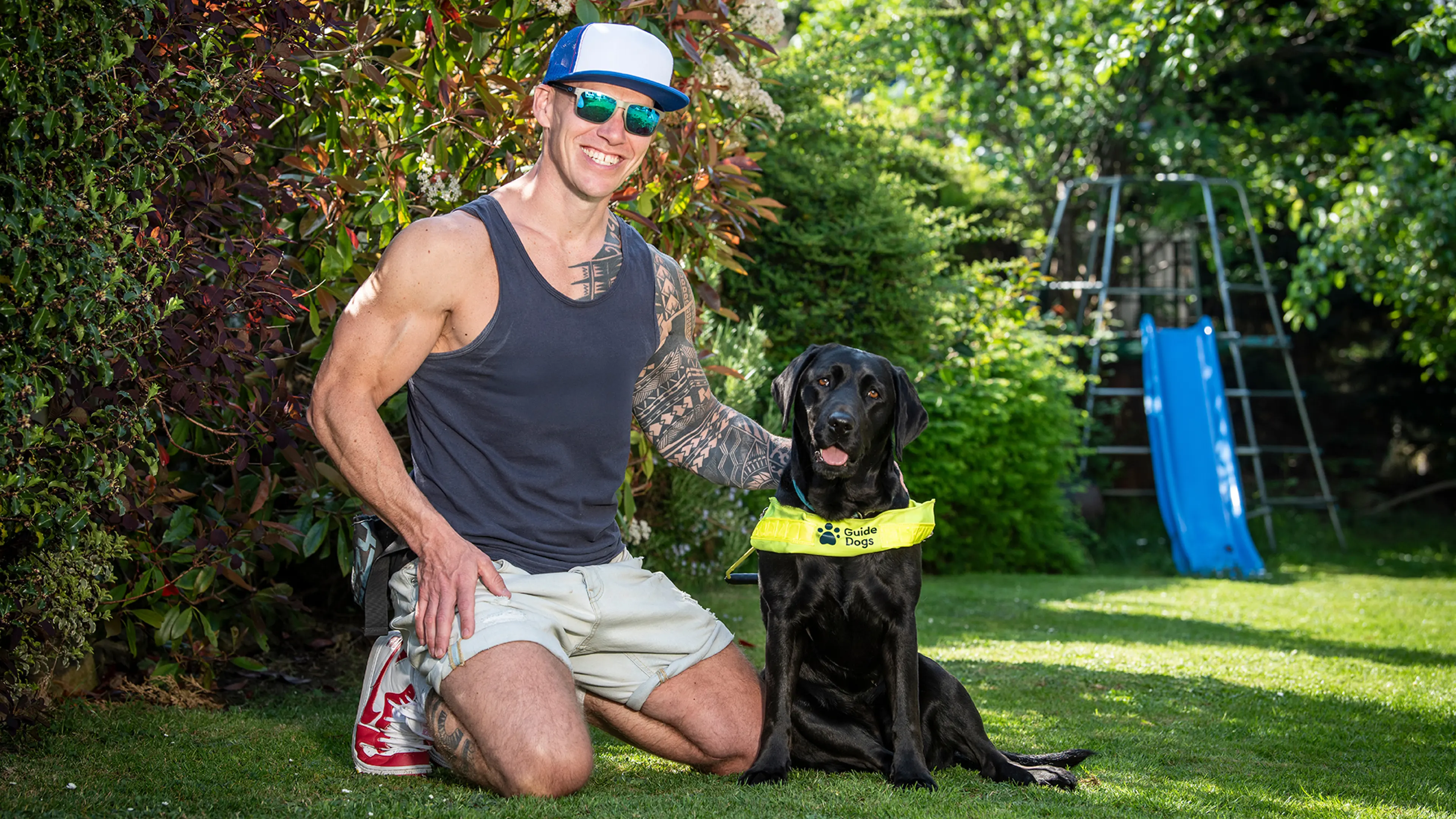 Guide dog owner Mark kneeling down next to black Labrador guide dog Mary outside in a garden.