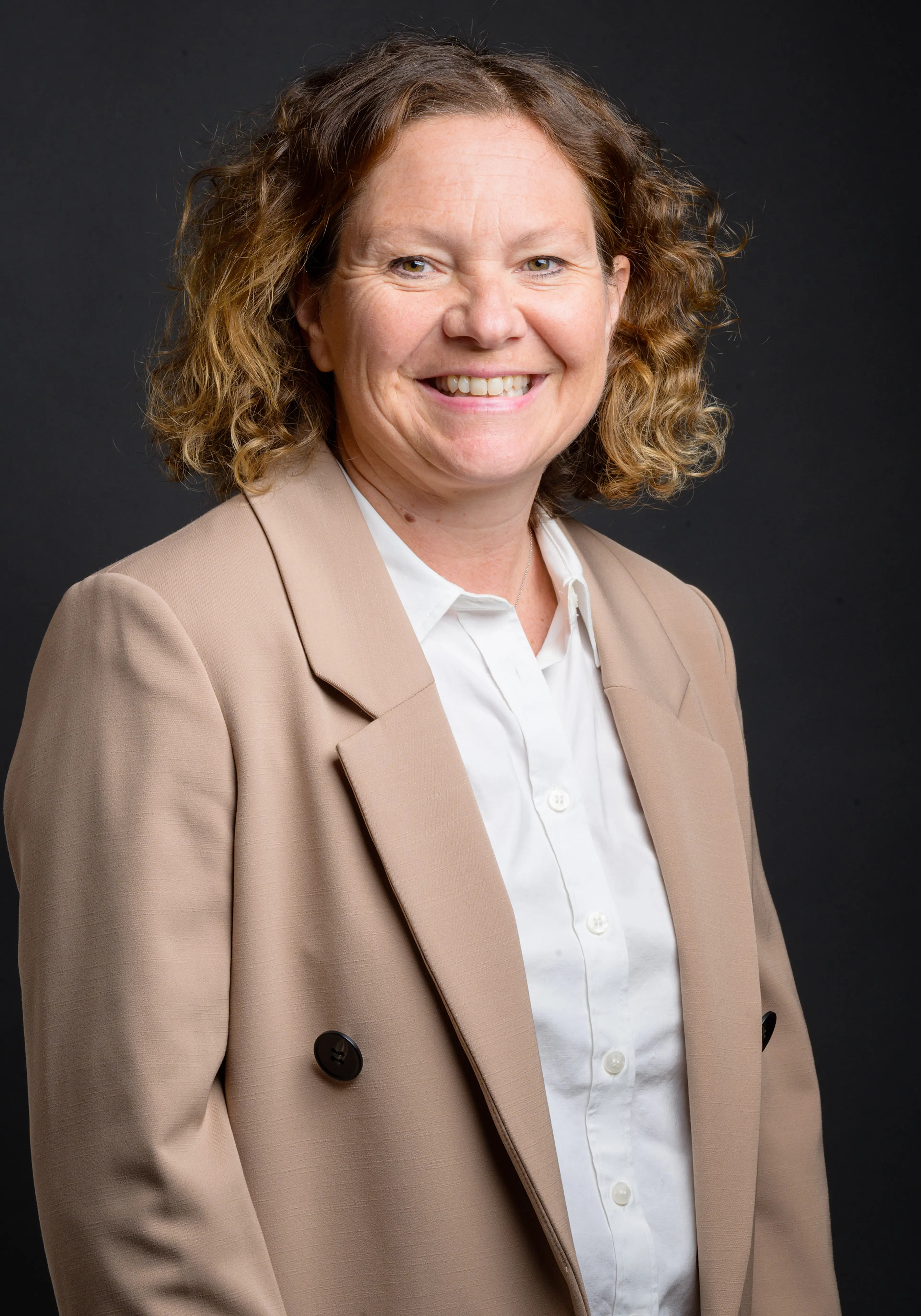 Kerry smiles at the camera. She has shoulder length brown curly hair and is wearing a beige suit.