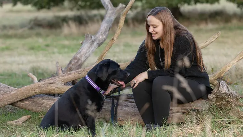 A dog owner sits on a fallen tree in a field, smiling at their dog, a black Labrador.