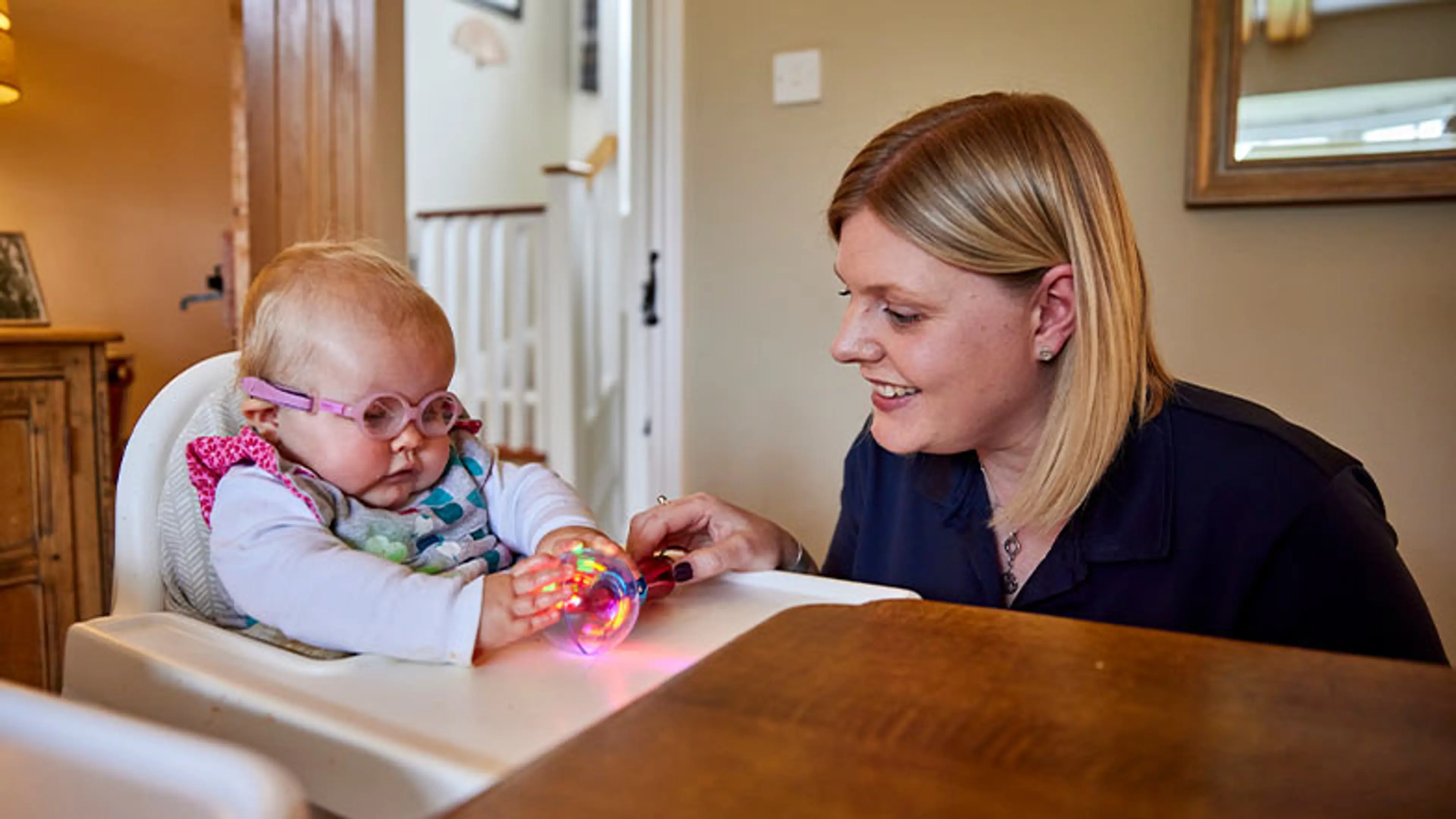 Margot, a baby who has a vision impairment, sits and plays in the living room with her Guide Dogs Habilitation Specialist, Kate.