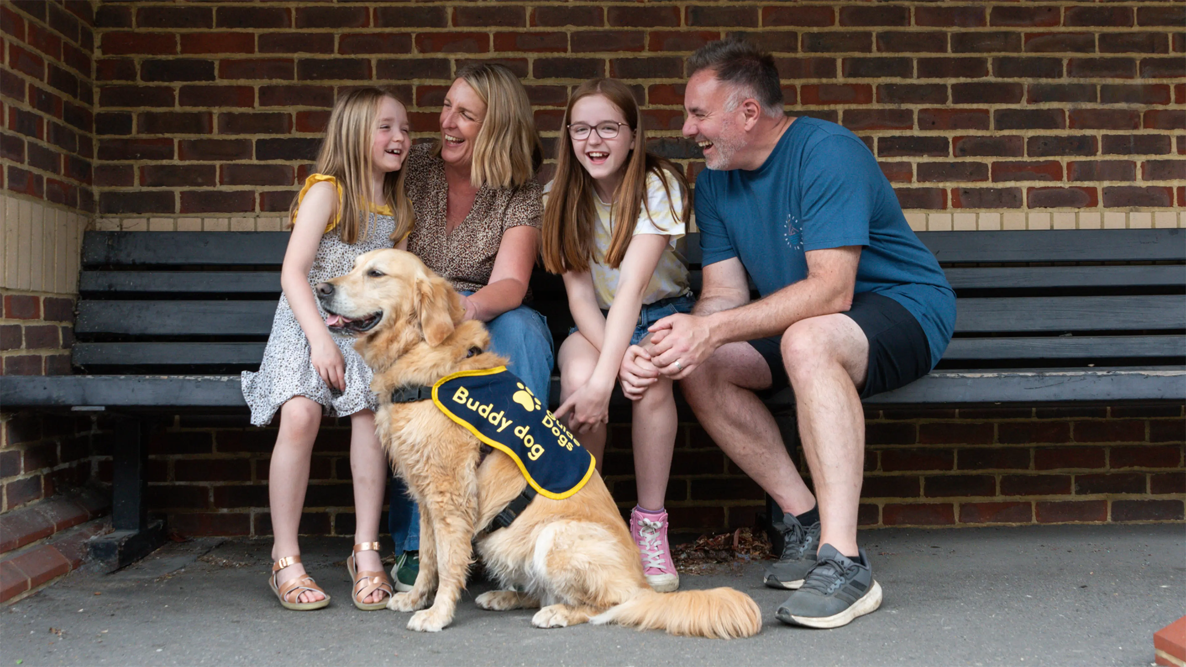 Freya, a young girl with a vision impairment, sits on a bench with her mum, dad and sister. Buddy dog Gwen sits at their feet.