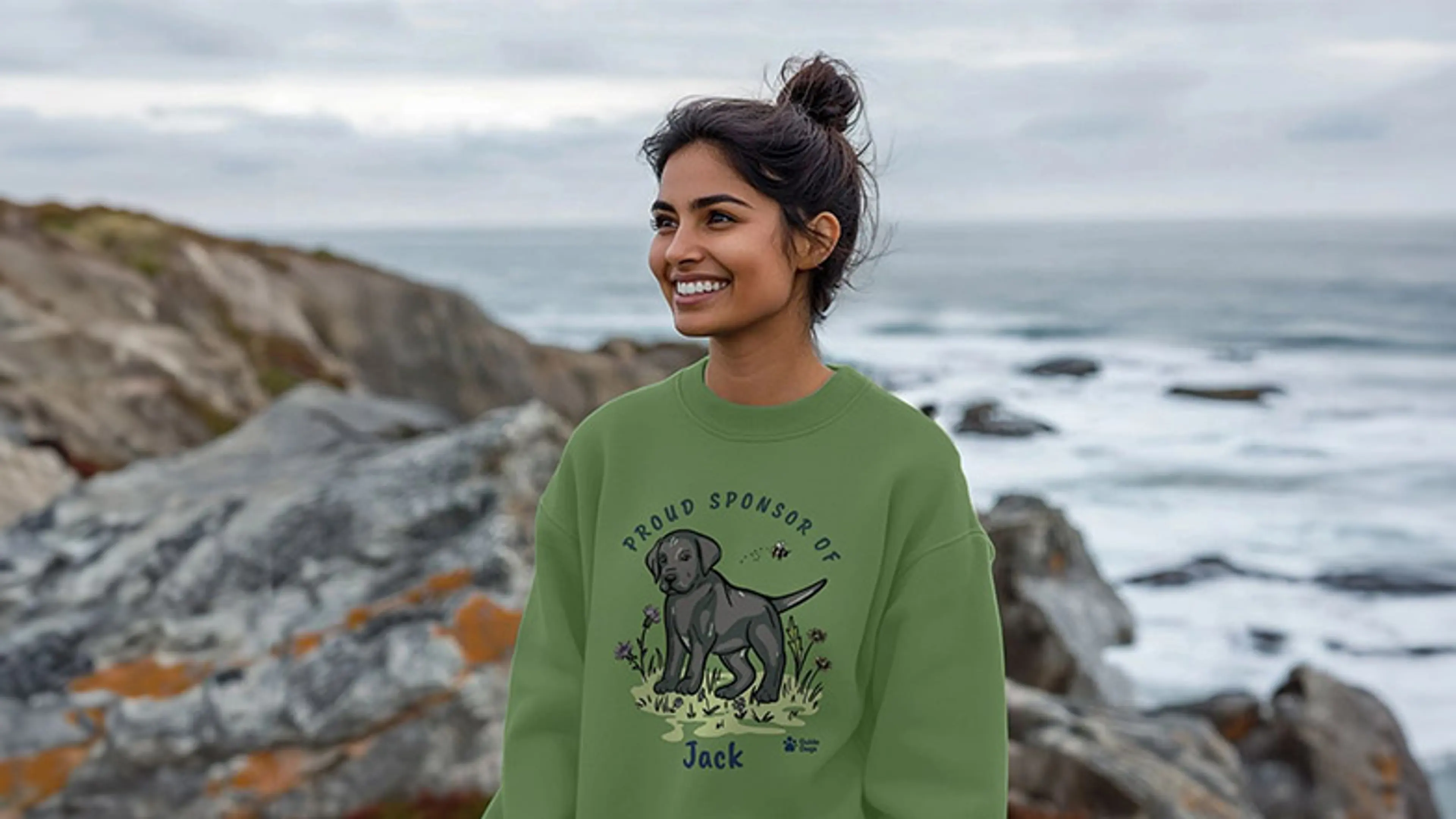 Smiling woman standing in front of a rocky outcrop with the sea in the background wearing a sweatshirt with sponsored pup Jack design.