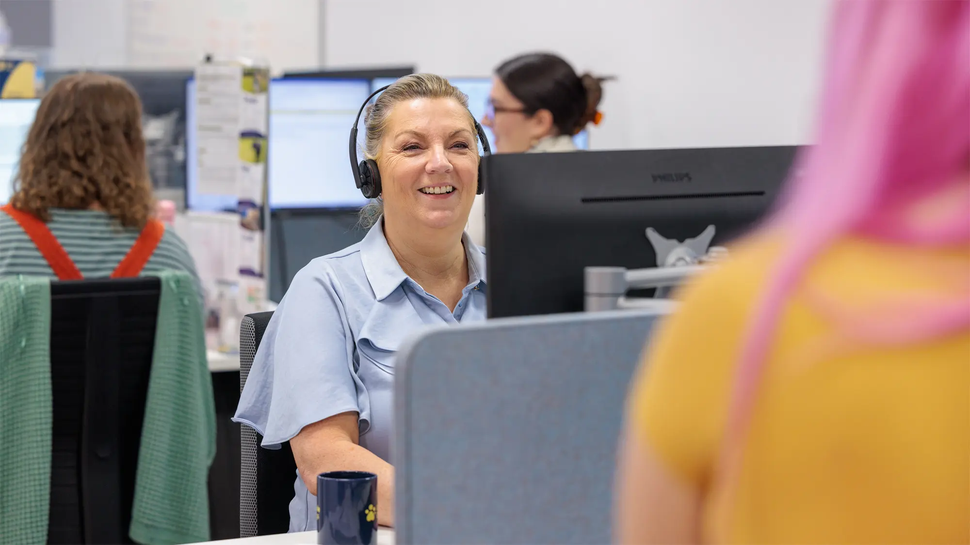 A Guide Dogs staff member wearing a headset smiles across her desk at another colleague.