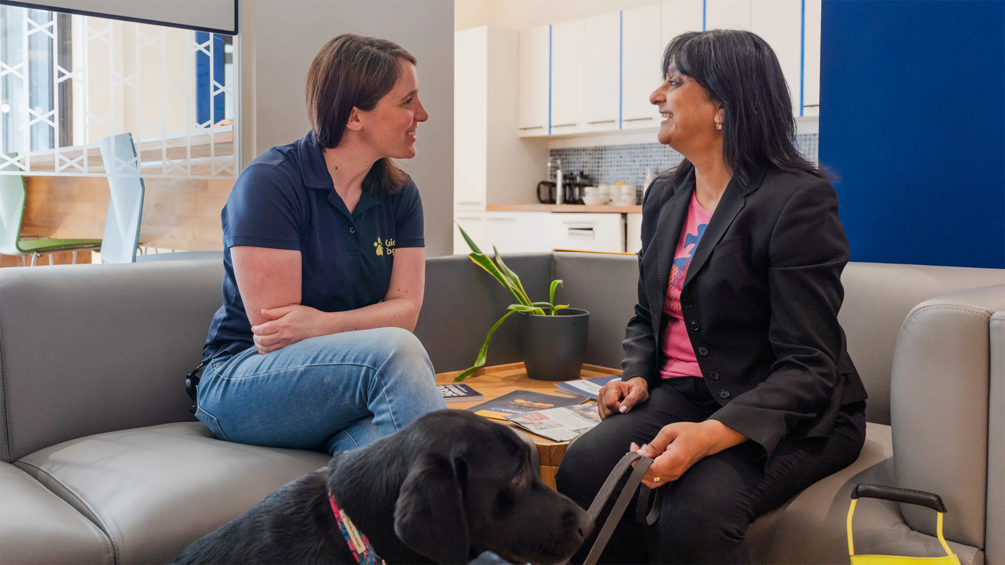 Deborah and her Guide Dogs Mobility Specialist Laura sit on a sofa in an office speaking. Deborah's black Labrador guide dog Betty sits in front of them.