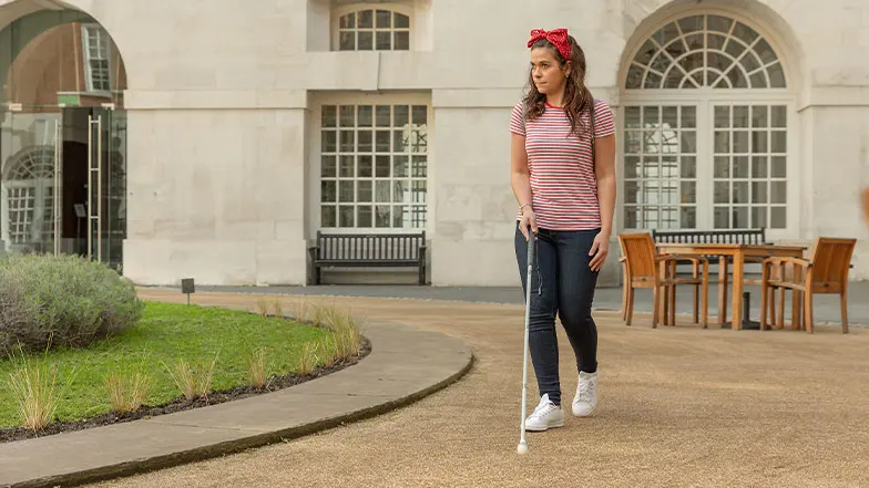 A woman with a red bow in her hair walks across a courtyard using her long cane to navigate.
