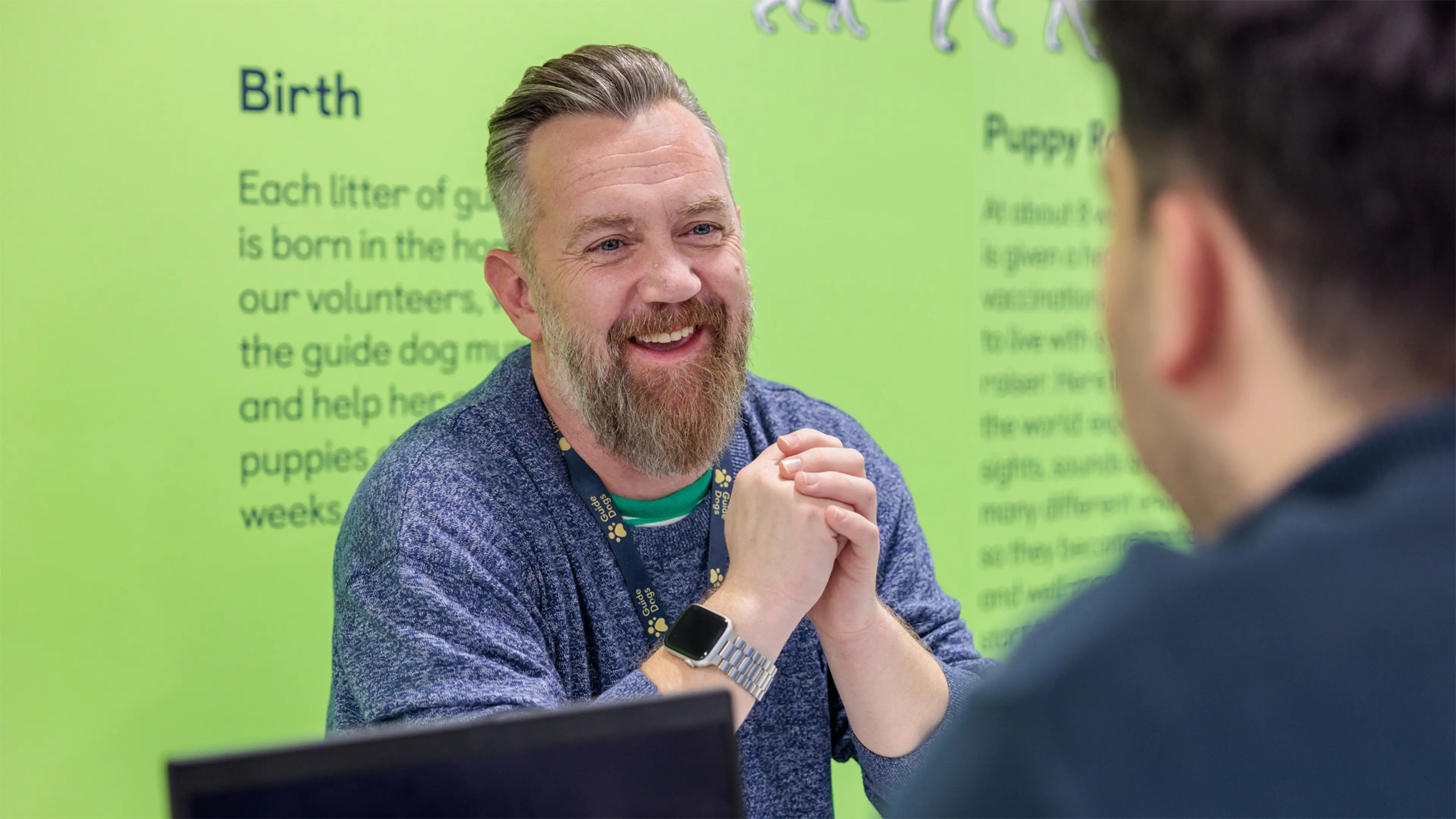 A Guide Dogs employee leans against a desk with his hands clasped whilst talking to a colleague.
