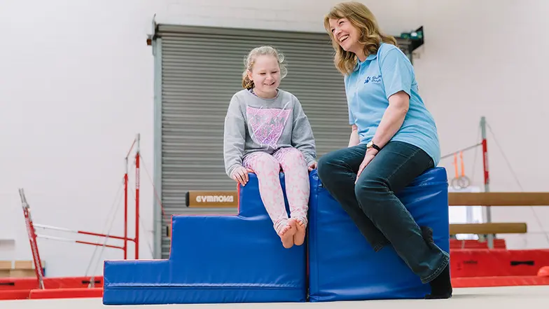 Oashia sitting on blue gymnastics block with Guide Dogs habilitation specialist