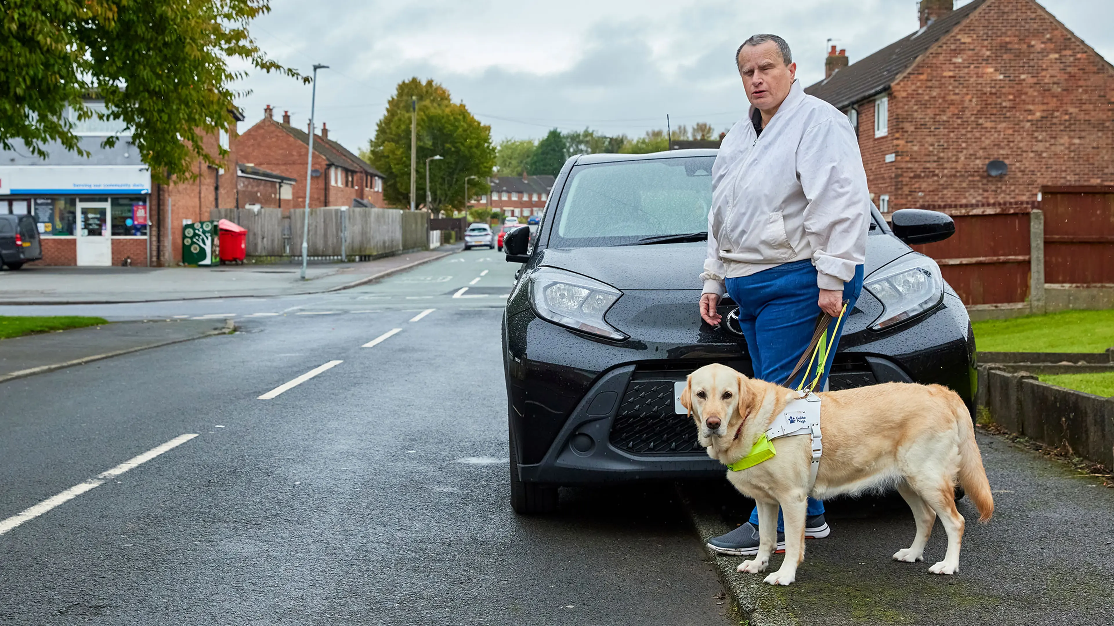 Guide dog owner Lee and his guide dog Millie stepping off the kerb into the road to pass a car that is parked on the pavement.