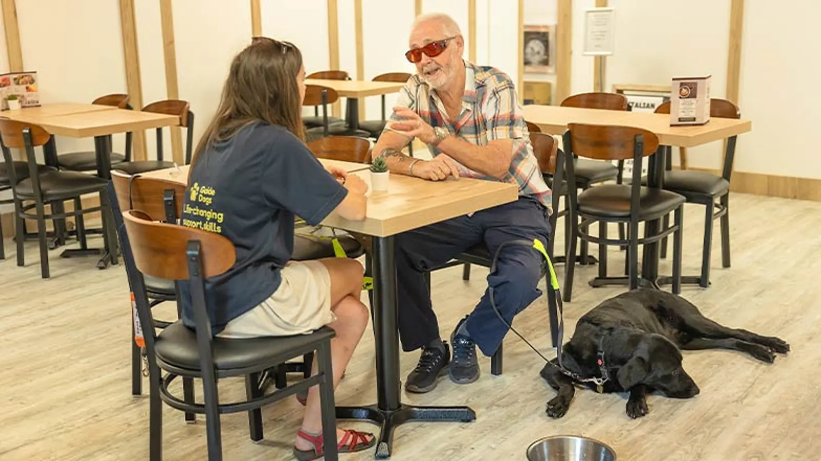 Guide dog owner Alan chats to a Guide Dog Mobility Specialist, in a cafe, with his guide dog, Dane, beside him.