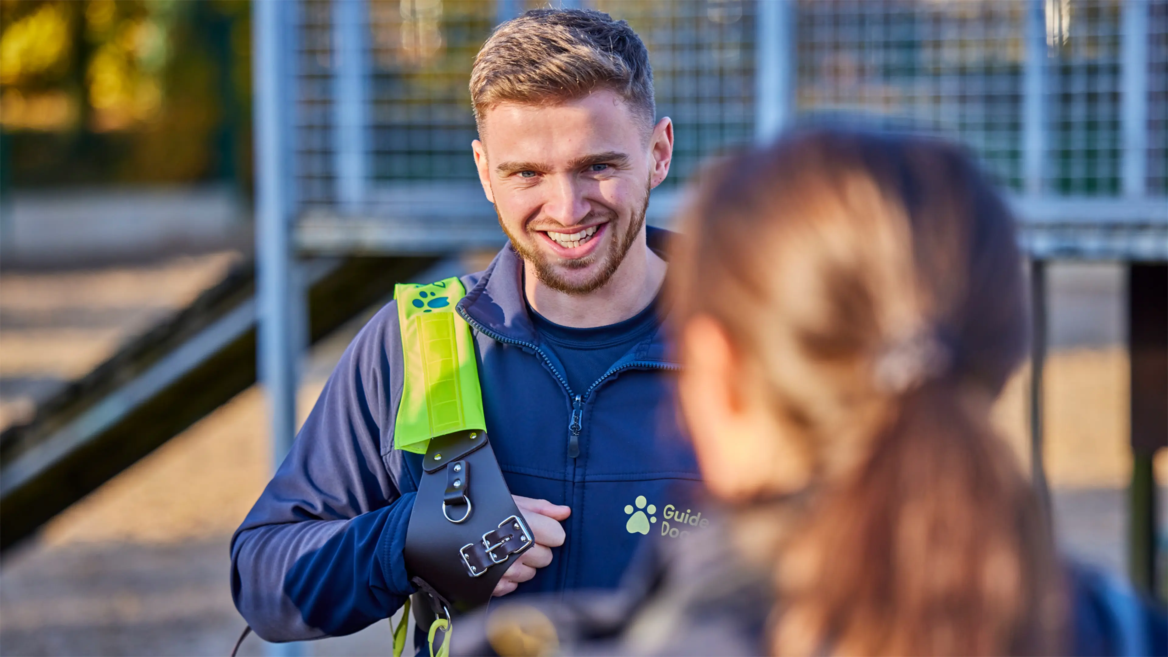 Two Guide Dogs Academy staff members stand talking outside. One has a guide dog harness resting on his shoulder.
