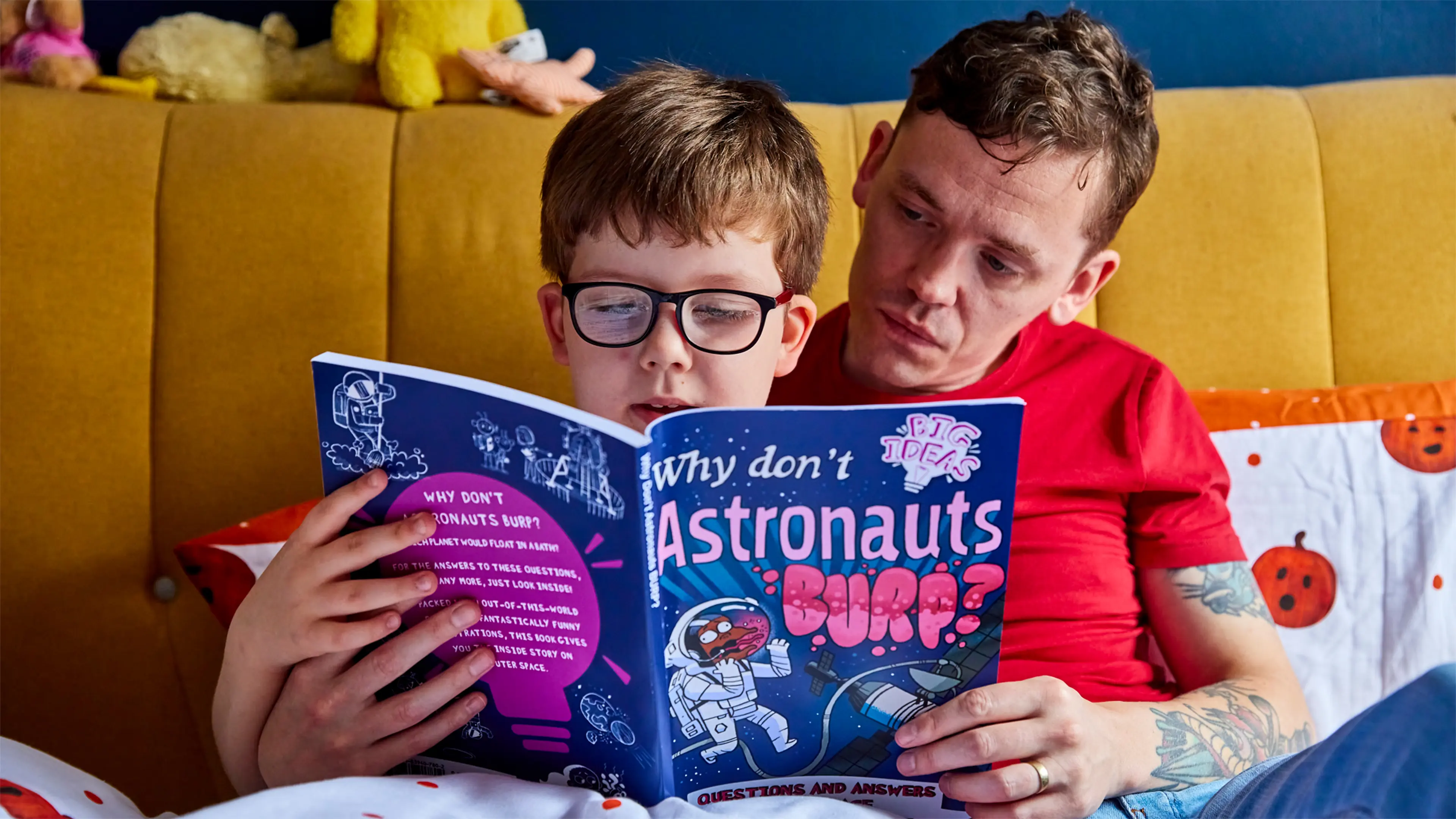 A boy with vision impairment and his dad sit on a bed together reading a large-print children's book.