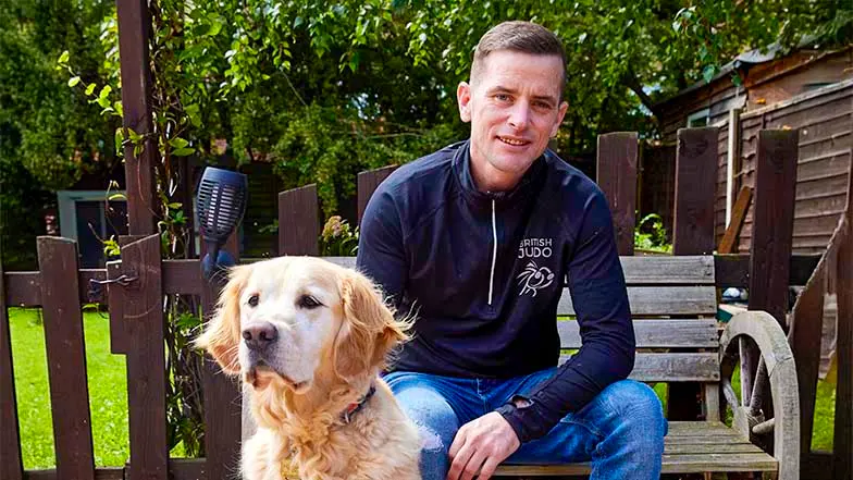 GDO Scott is sitting on a bench, looking to the camera with guide dog Milo in front of him.