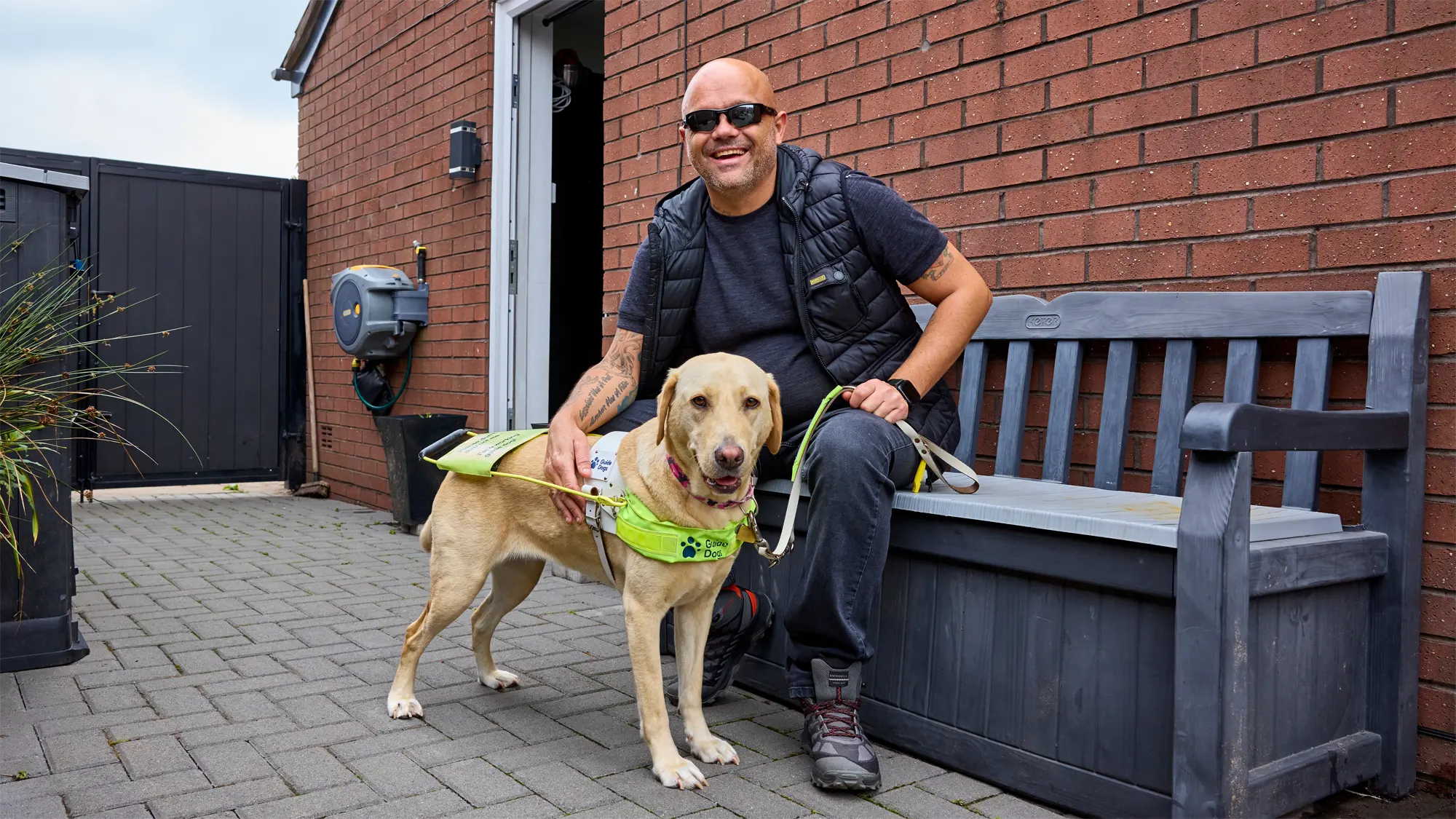 Guide dog owner Wayne sits on a bench in his garden. His yellow Labrador Liberty stands beside him.