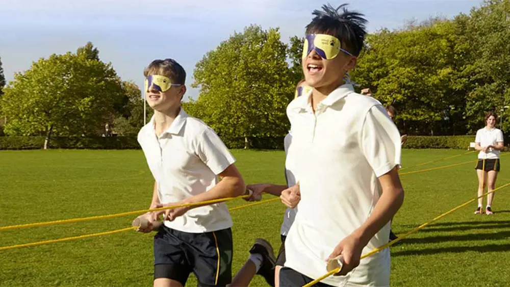 Teenage school children running a race in sports day blindfolded and using a rope to guide them.