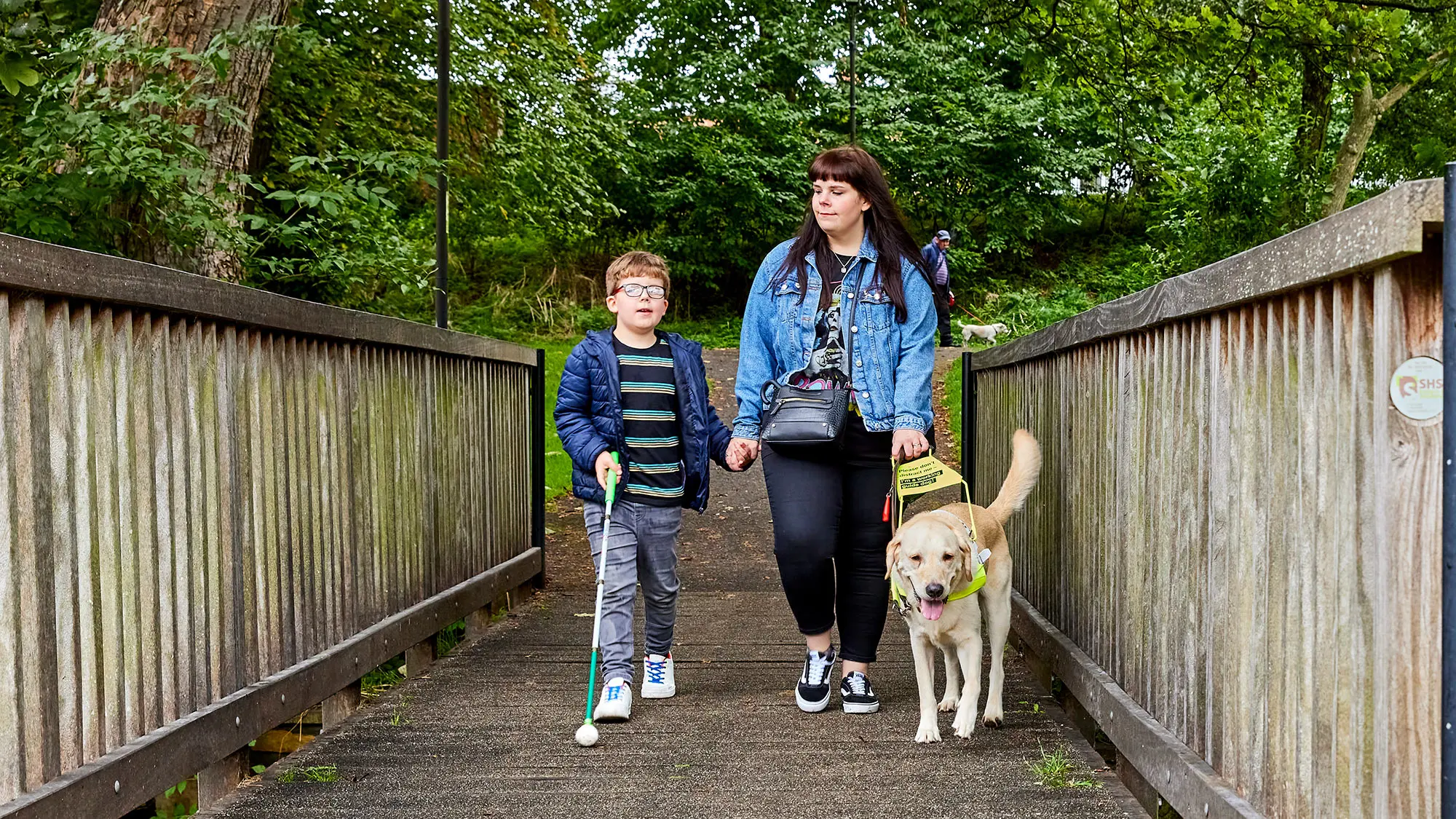 Guide Dog owner Lorna walks across a wooden bridge with her guide dog and son who is using a cane. 