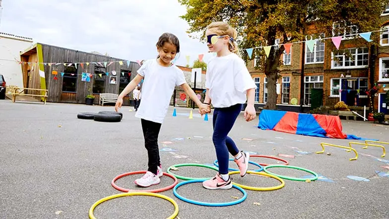 Two schoolchildren doing an obstacle race, one blindfolded and one holding their hand to guide them.