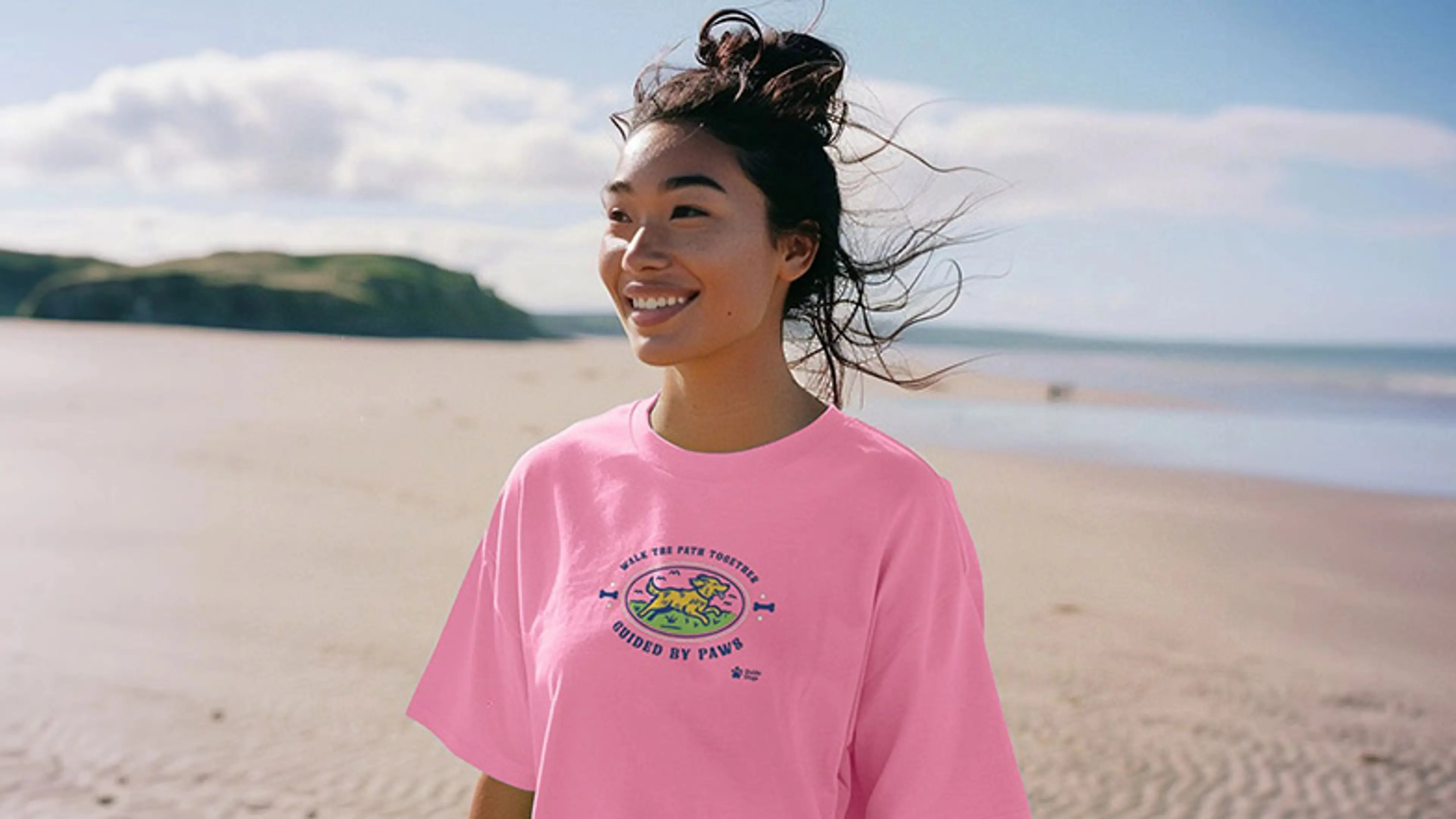 A woman is at the beach wearing a pink t-shirt from the Guide Dogs Teemill shop