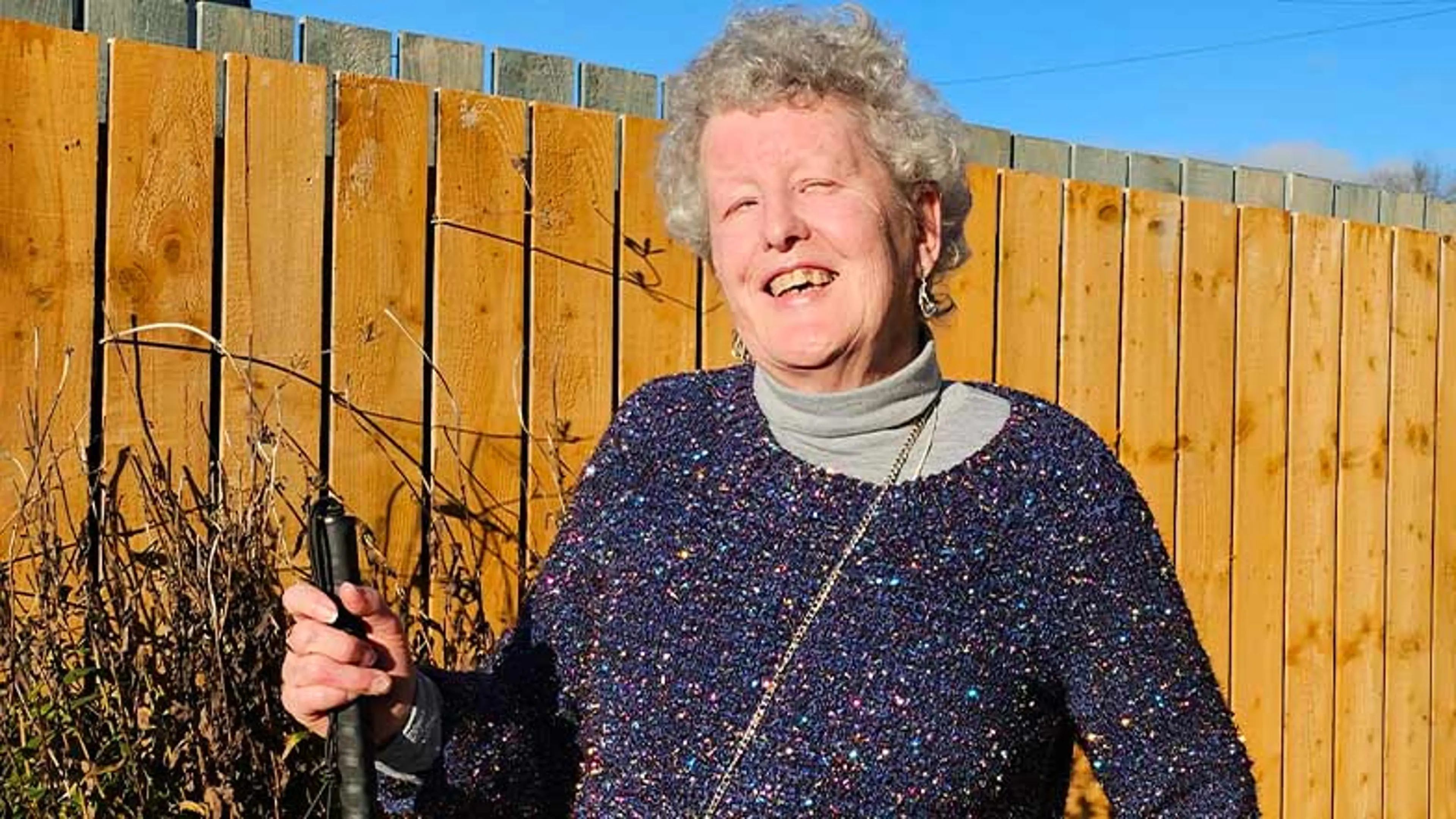 Margaret stands smiling in the sunshine, holding a long cane.