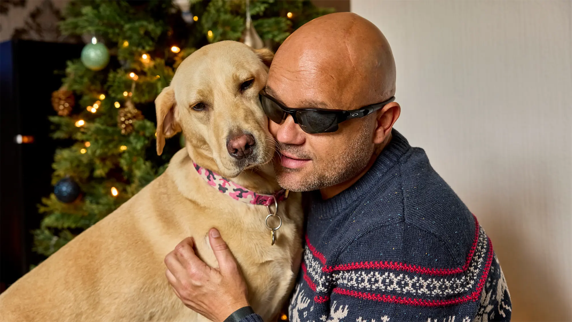 Guide dog owner Wayne wears a festive jumper and cuddles his Labrador guide dog Liberty in front of a Christmas tree.