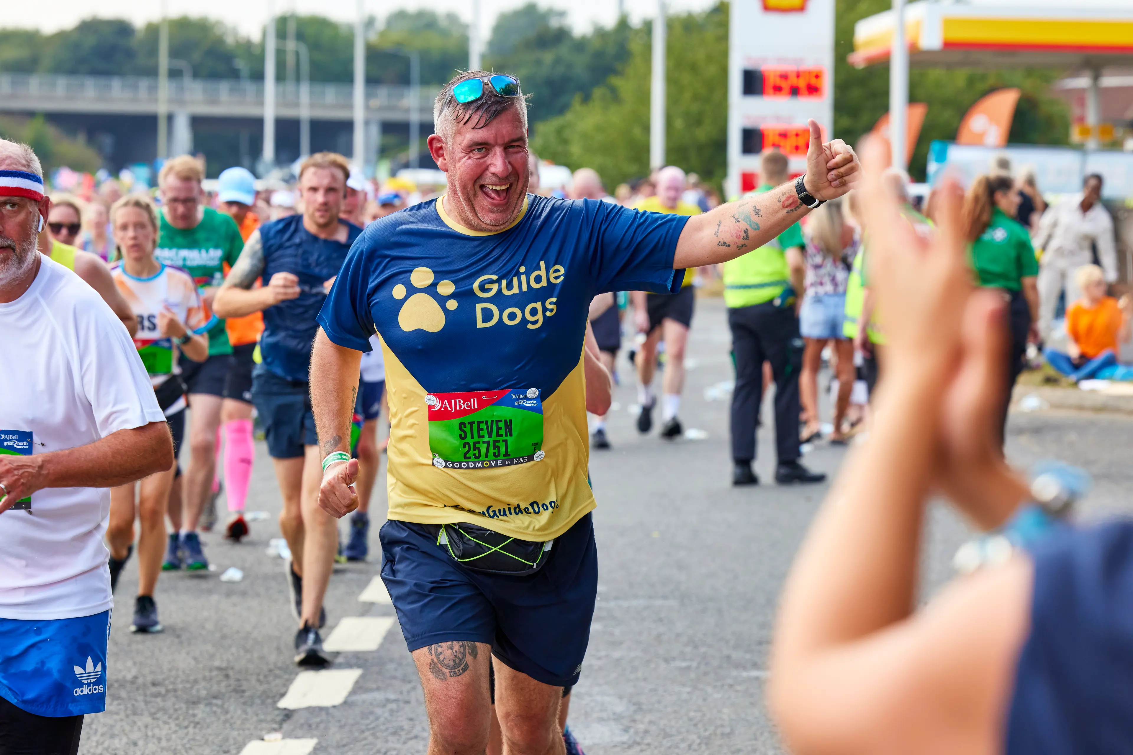 A runner for Guide Dogs waves his hand in the air giving a thumbs up as he passes cheering supporters.