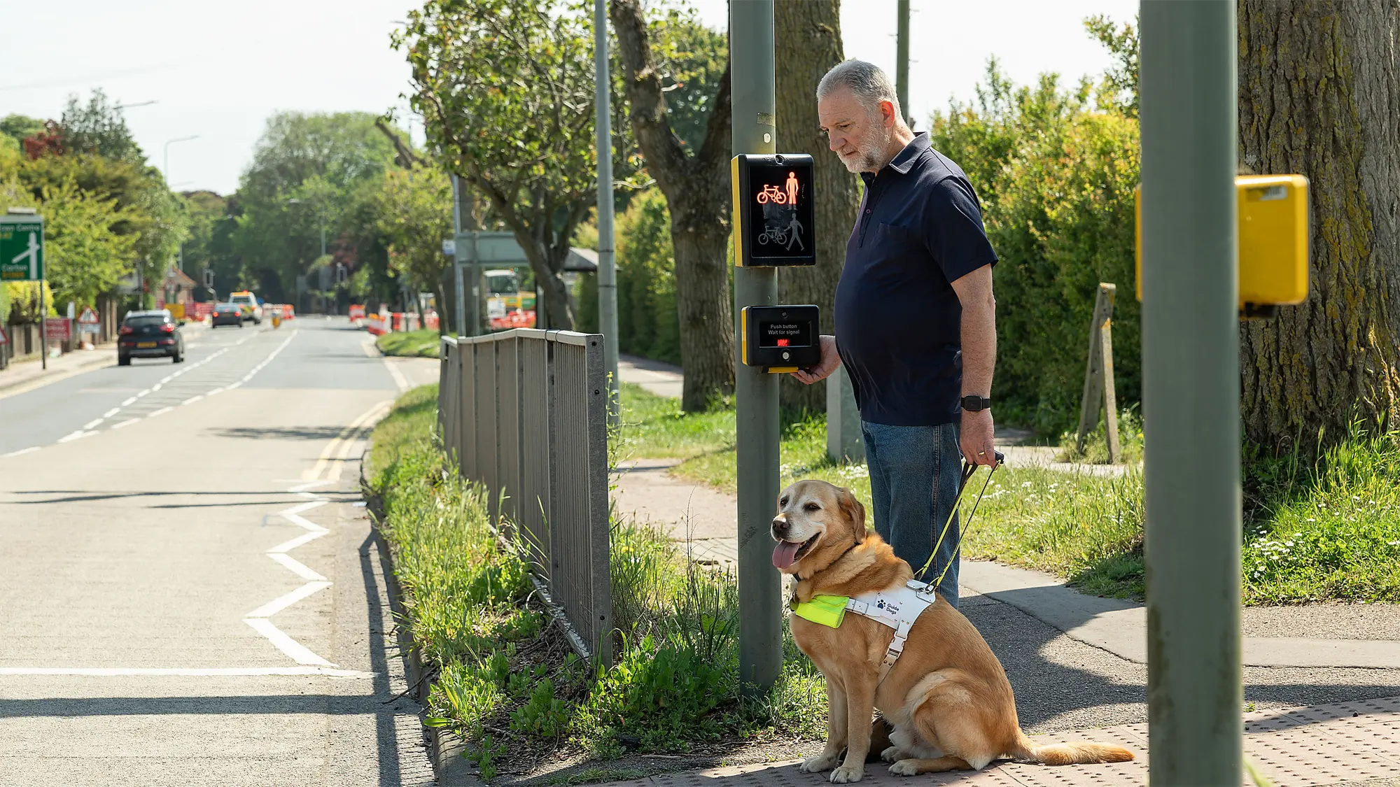 Guide dog owner Simon waits at a pedestrian crossing with one hand on the tactile crossing cone, with his guide dog Mayne.