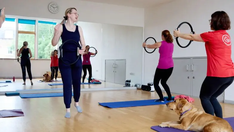 Emma stands in an exercise studio teaching a pilates class and holding a pilates ring.