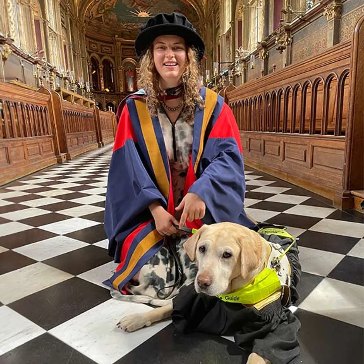 A smiling Anica in her graduation robes with guide dog Lassie at her feet.