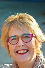 Lynn smiles at the camera. She has short blonde hair and is wearing colourful glasses.