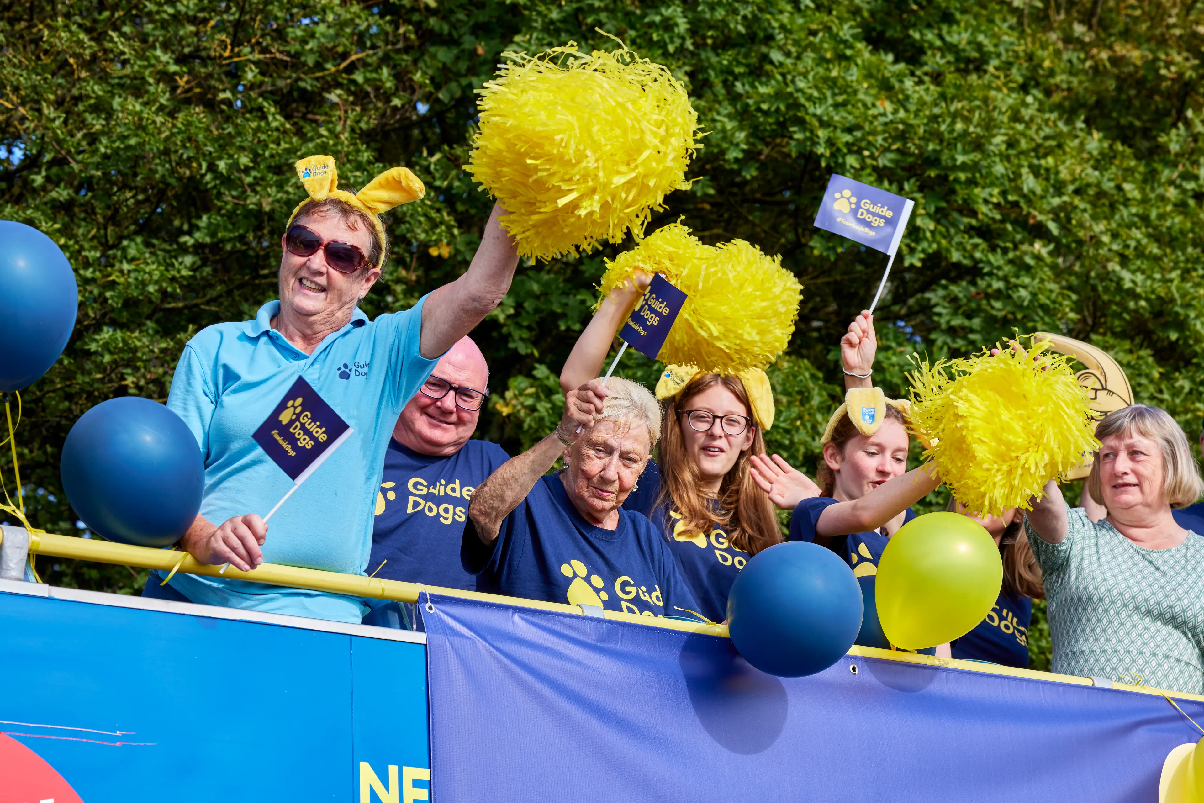 Open-top double decker bus filled with cheering supporters. The bus has a Guide Dogs banner and blue and yellow balloons.