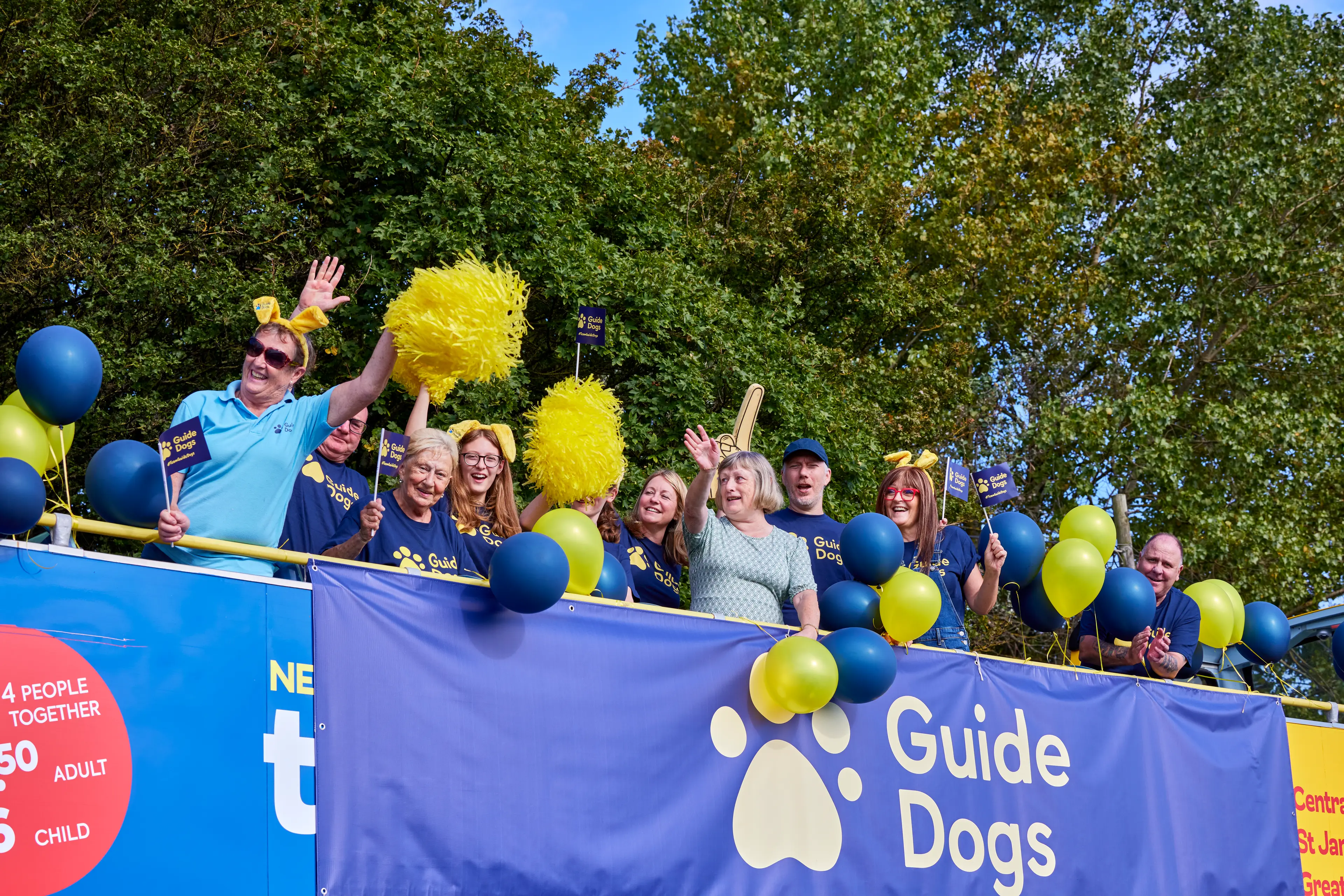 Open-top double decker bus filled with cheering supporters. The bus has a Guide Dogs banner and blue and yellow balloons.