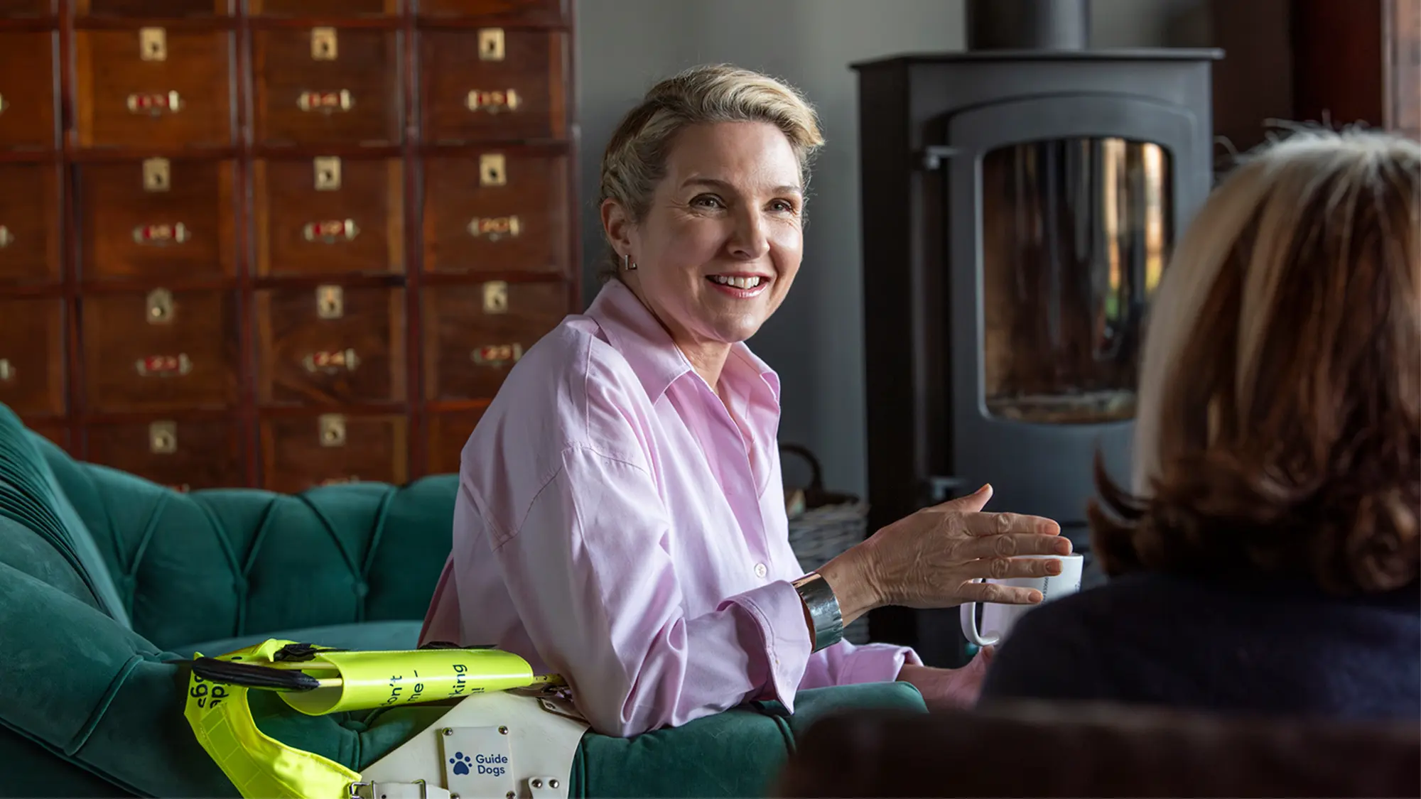 Guide dog owner and dressage rider Verity sits on an armchair, her guide dog's harness rests on the armchair.