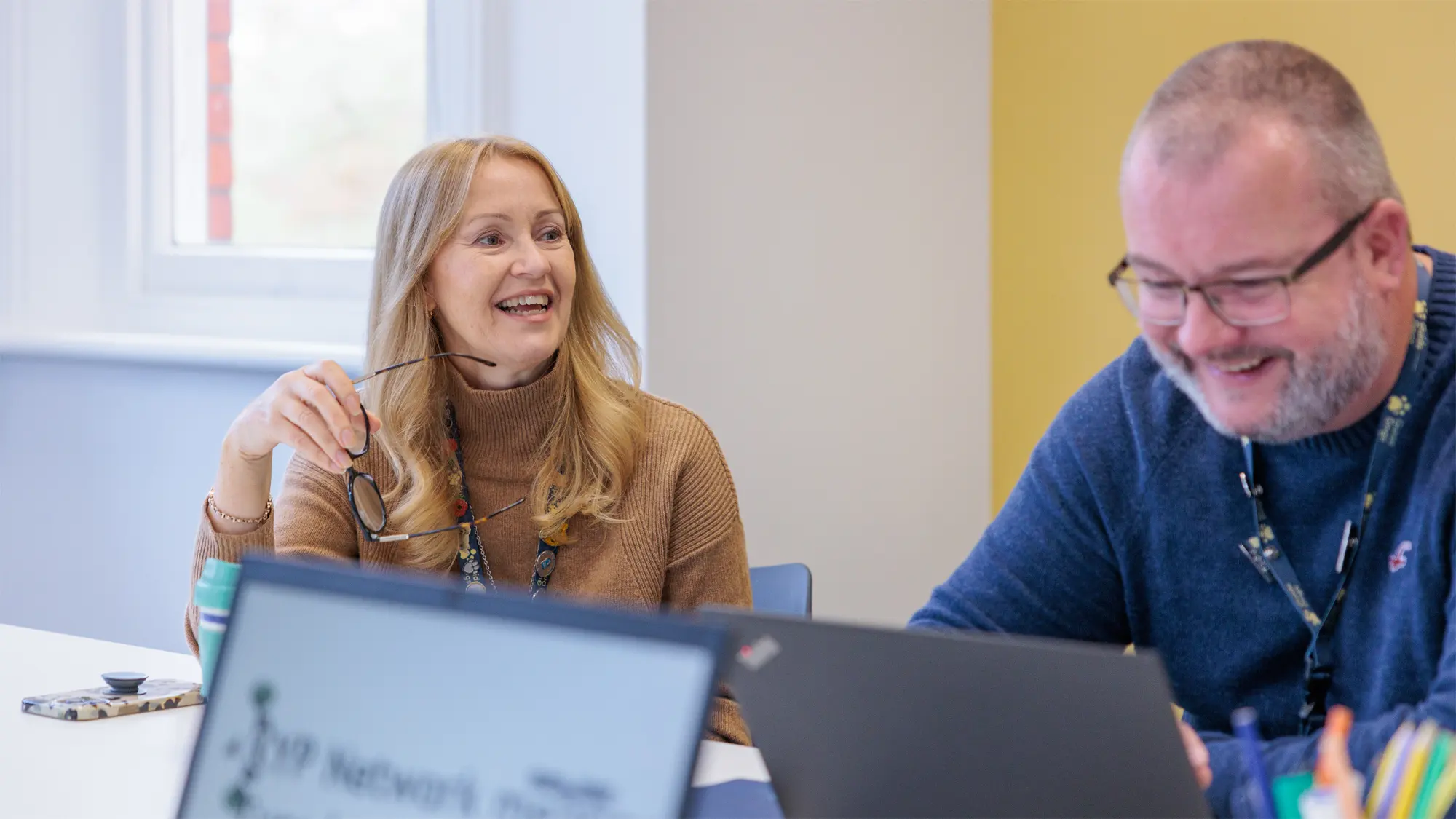 Two members of Guide Dogs staff laugh and smile as they sit at their laptops in the office.