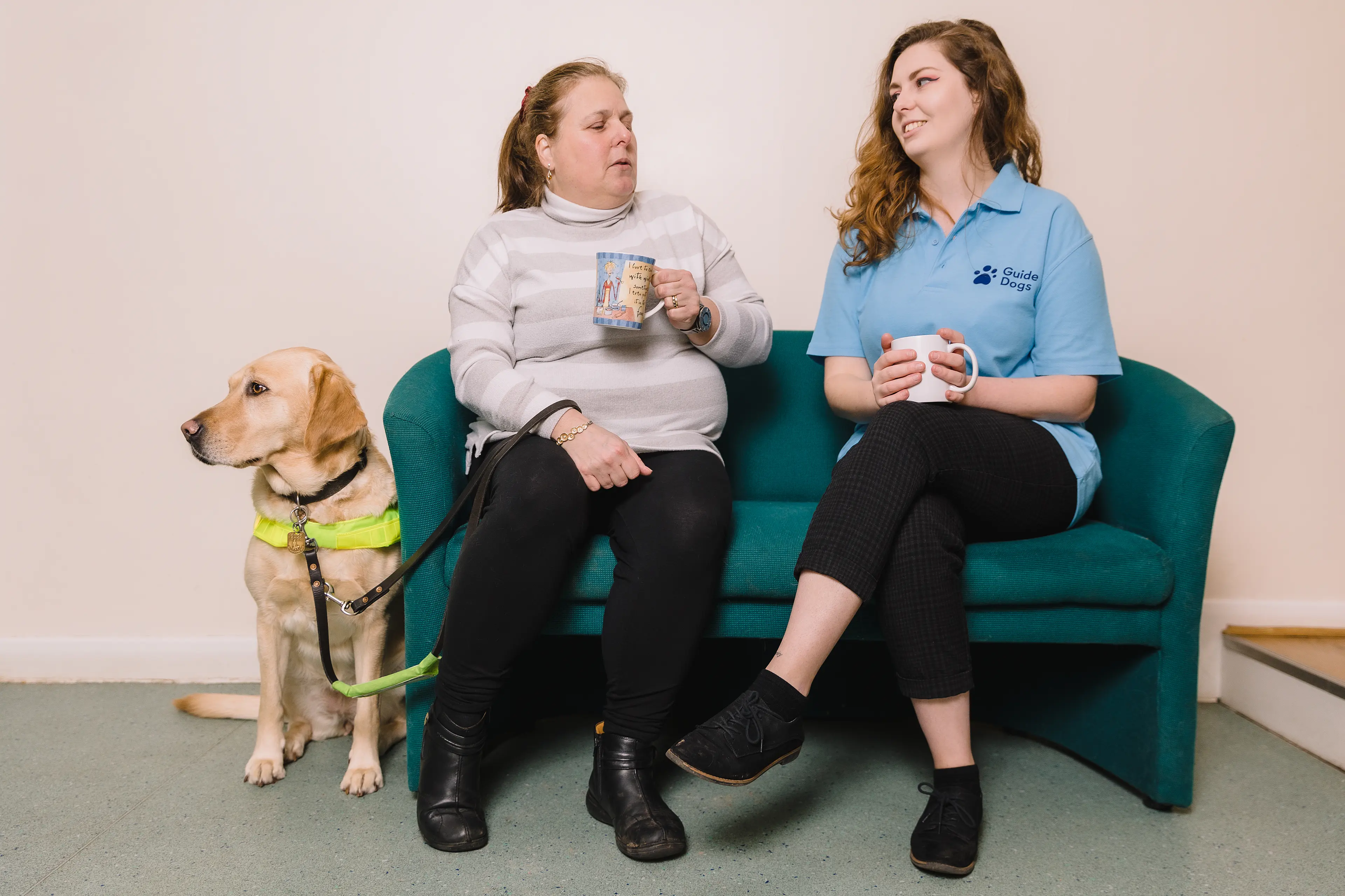 Two ladies sit side by side on a sofa while a guide dog in harness sits beside them.