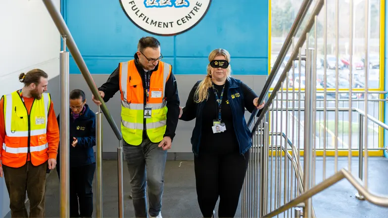 A Guide Dogs member of staff wears a blindfold and is led up the stairs by an Amazon staff member, as they practice sighted guiding.