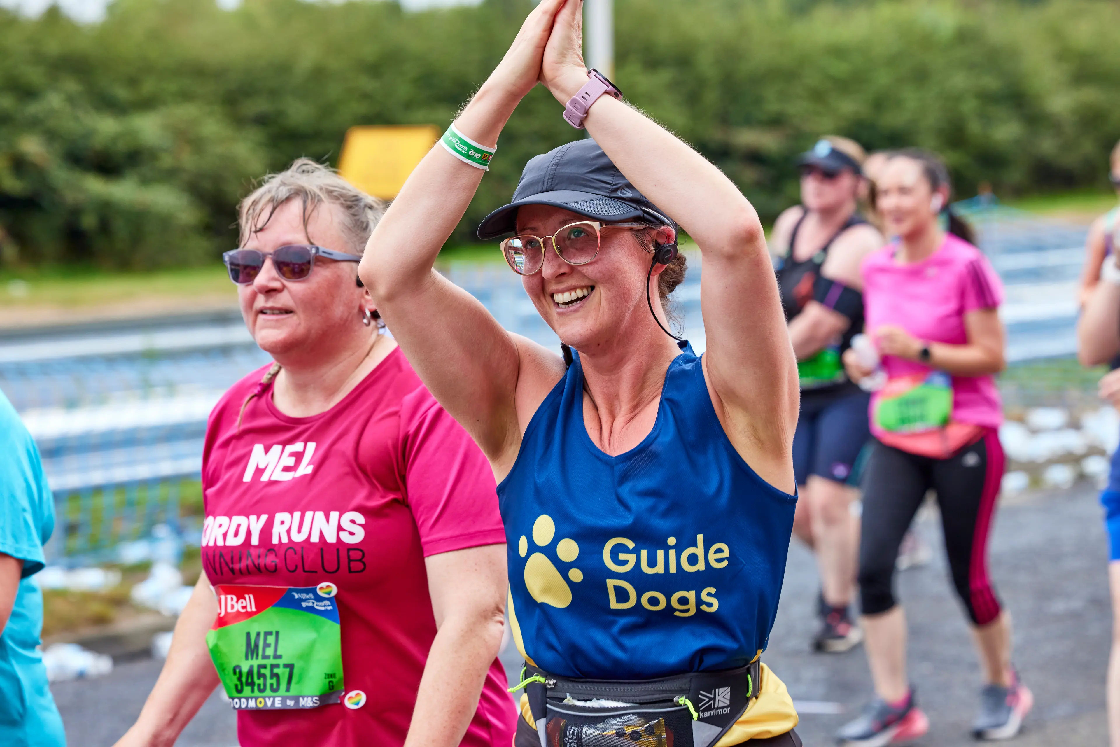 A runner in a race wearing a Guide Dogs vest smiles at the camera and waves her arms in the air.