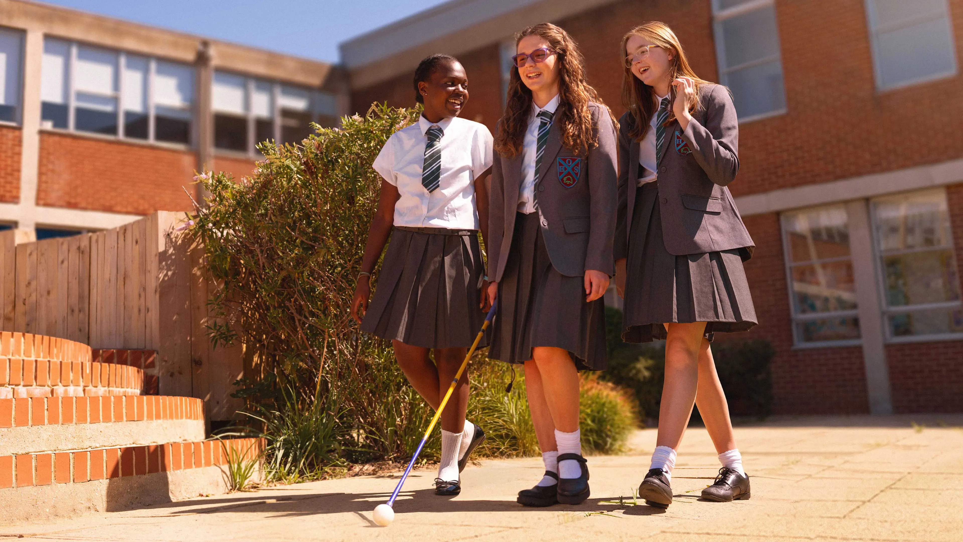 Erin and her two friends walk along a path in their school uniforms. Erin is using her long cane.