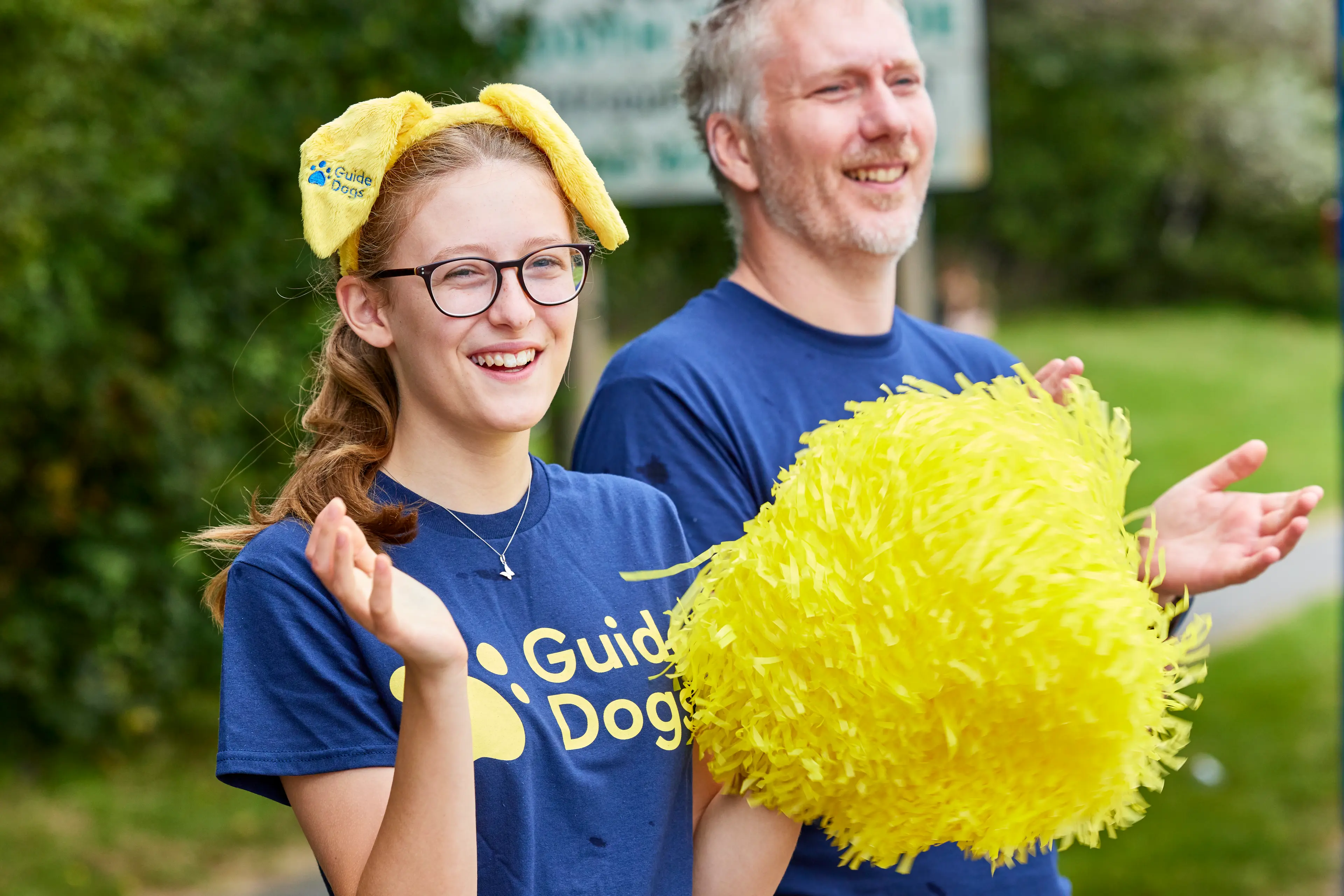 A girl and man in the crowd cheering at a race. They are wearing Guide Dogs t-shirts and ears.