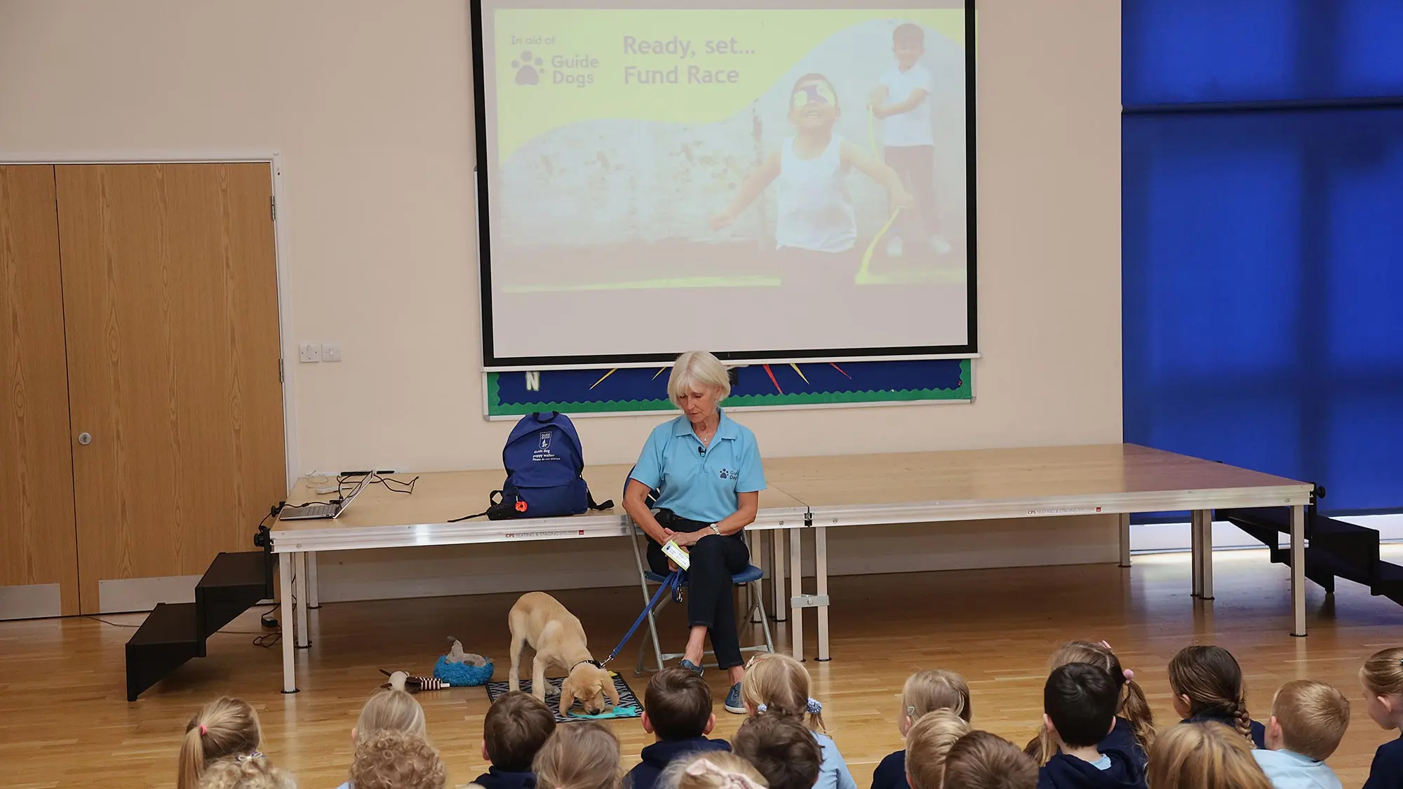 A Guide Dogs speaker with their dog at a school assembly with a Fund Race presentation behind them.