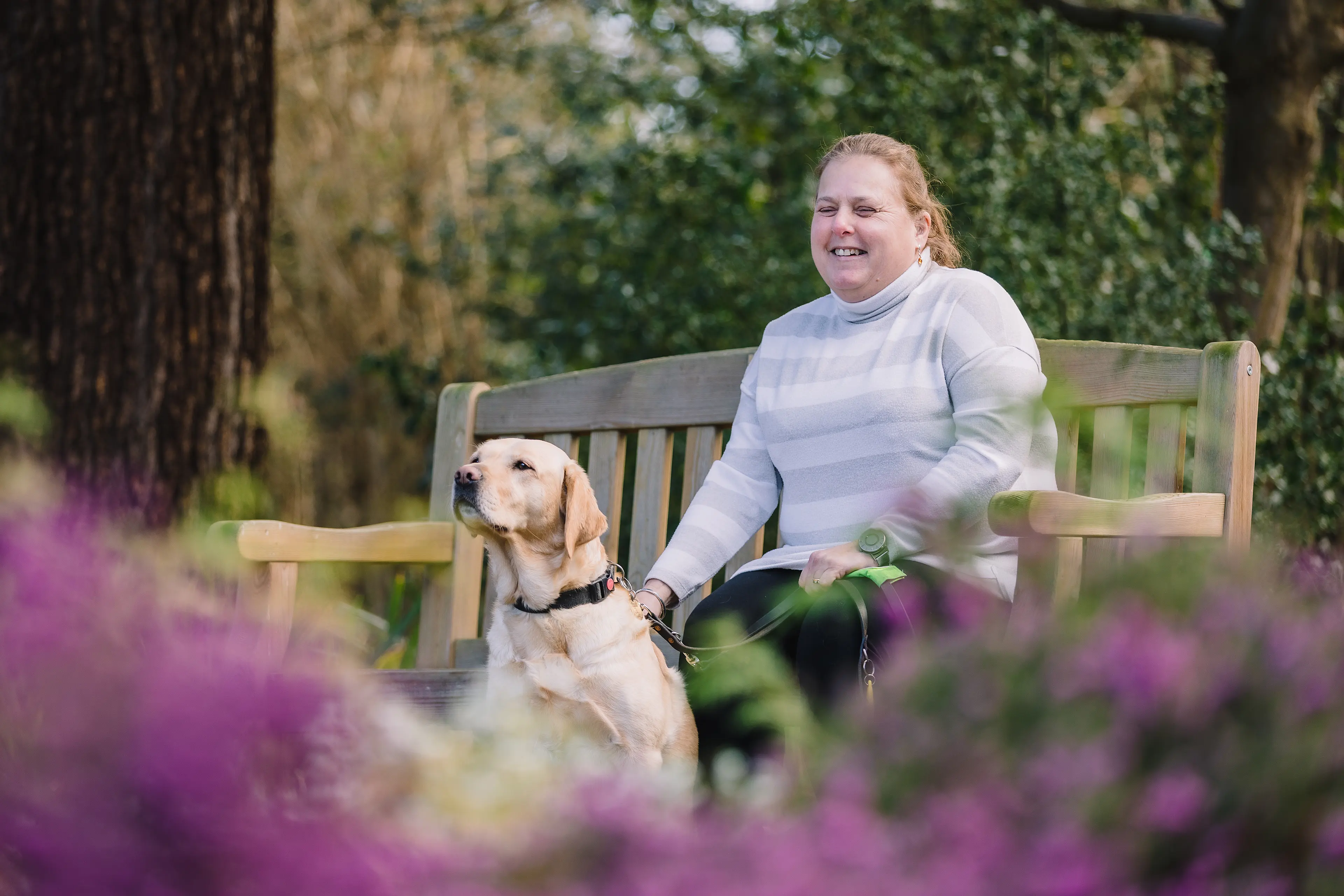 A lady sits on a bench with her guide dog by her side.