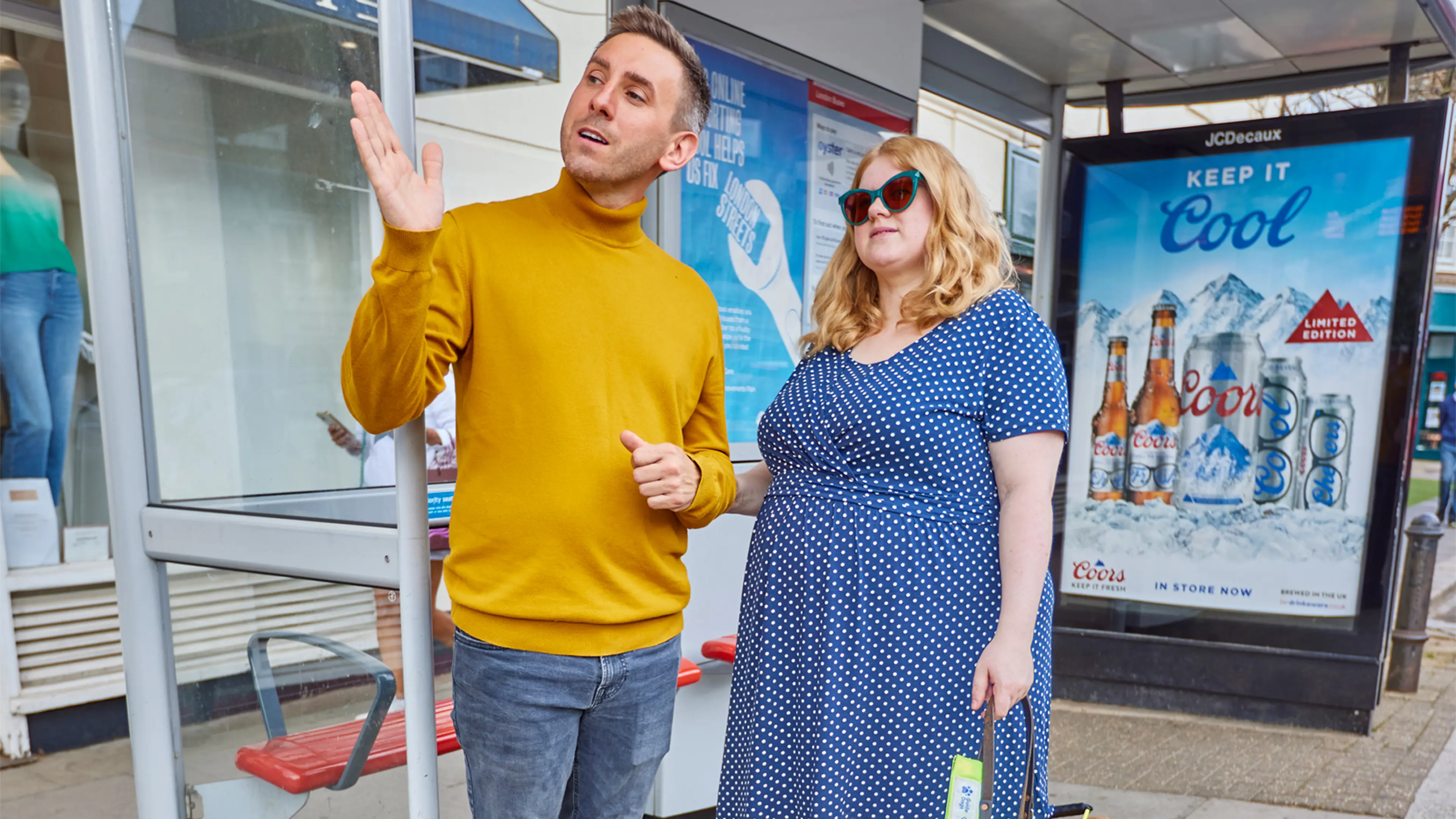 A guide dog owner stands at a bus stop with her golden retriever guide dog and a sighted guide, who is explaining what's around her.